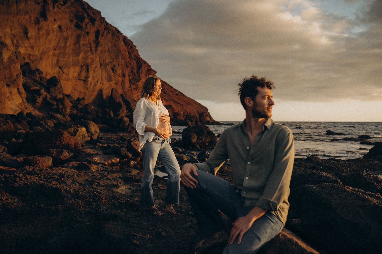 A couple poses on rocky coastal cliffs at golden hour, with dramatic ocean waves and warm sunlit rock formations behind them.
