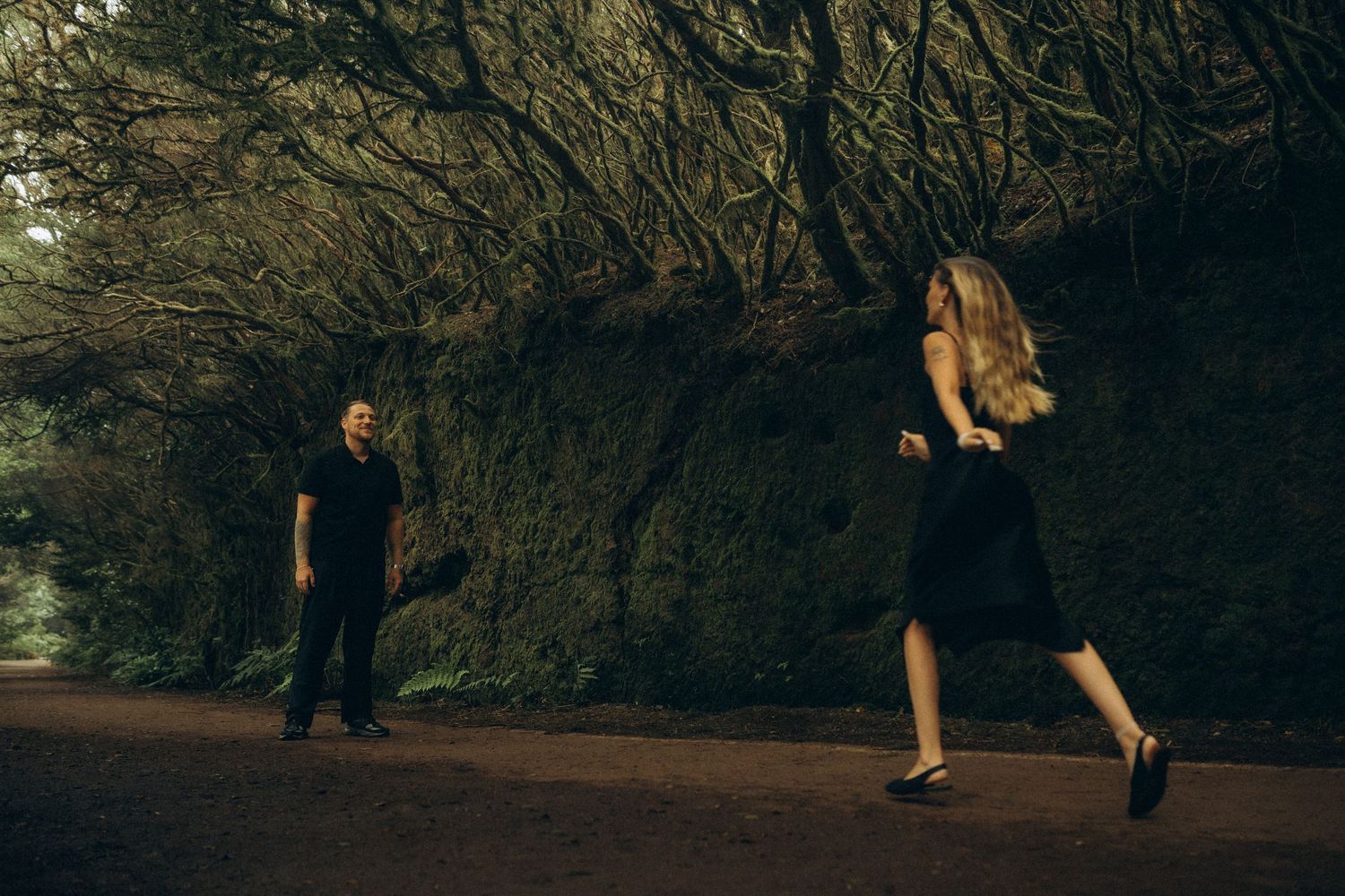 A man and woman in black outfits walk on a dark forest path surrounded by moss-covered walls and dense trees.