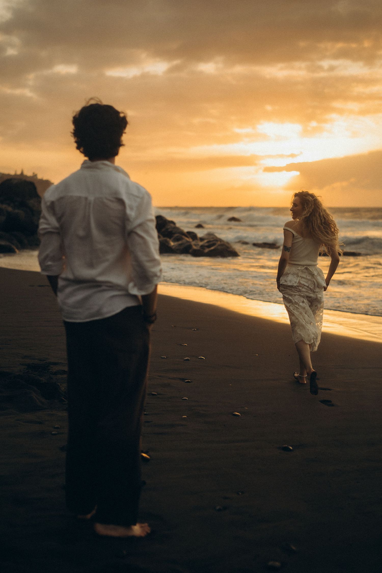 Couple on a black sand beach at sunset, woman in white dress walking toward ocean waves as man watches from behind.