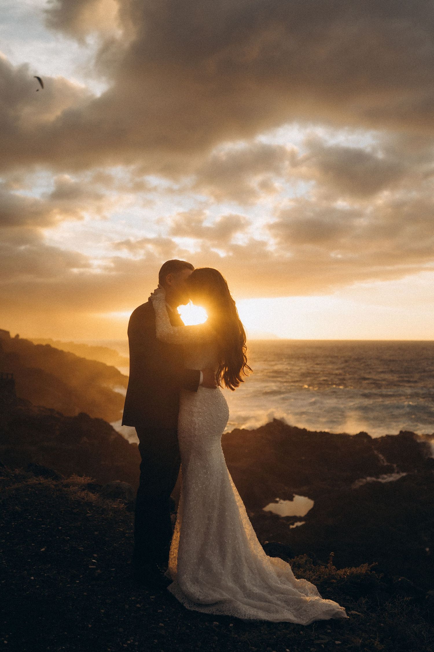 Wedding couple standing in embrace with sunset between them, Costa El Sauzal