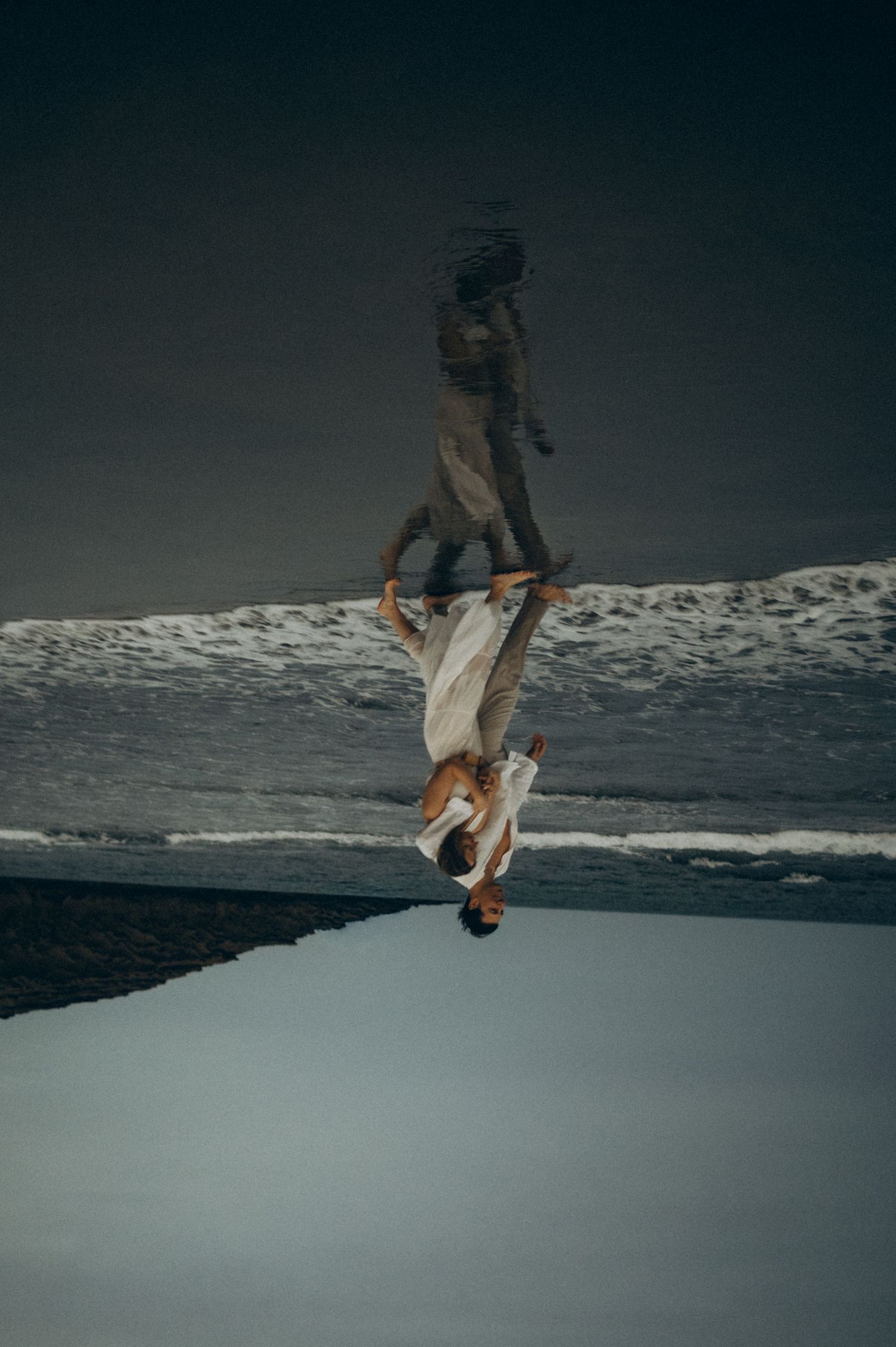 A surreal reflection of a woman in white dress on wet beach sand, creating a mirrored double image effect.