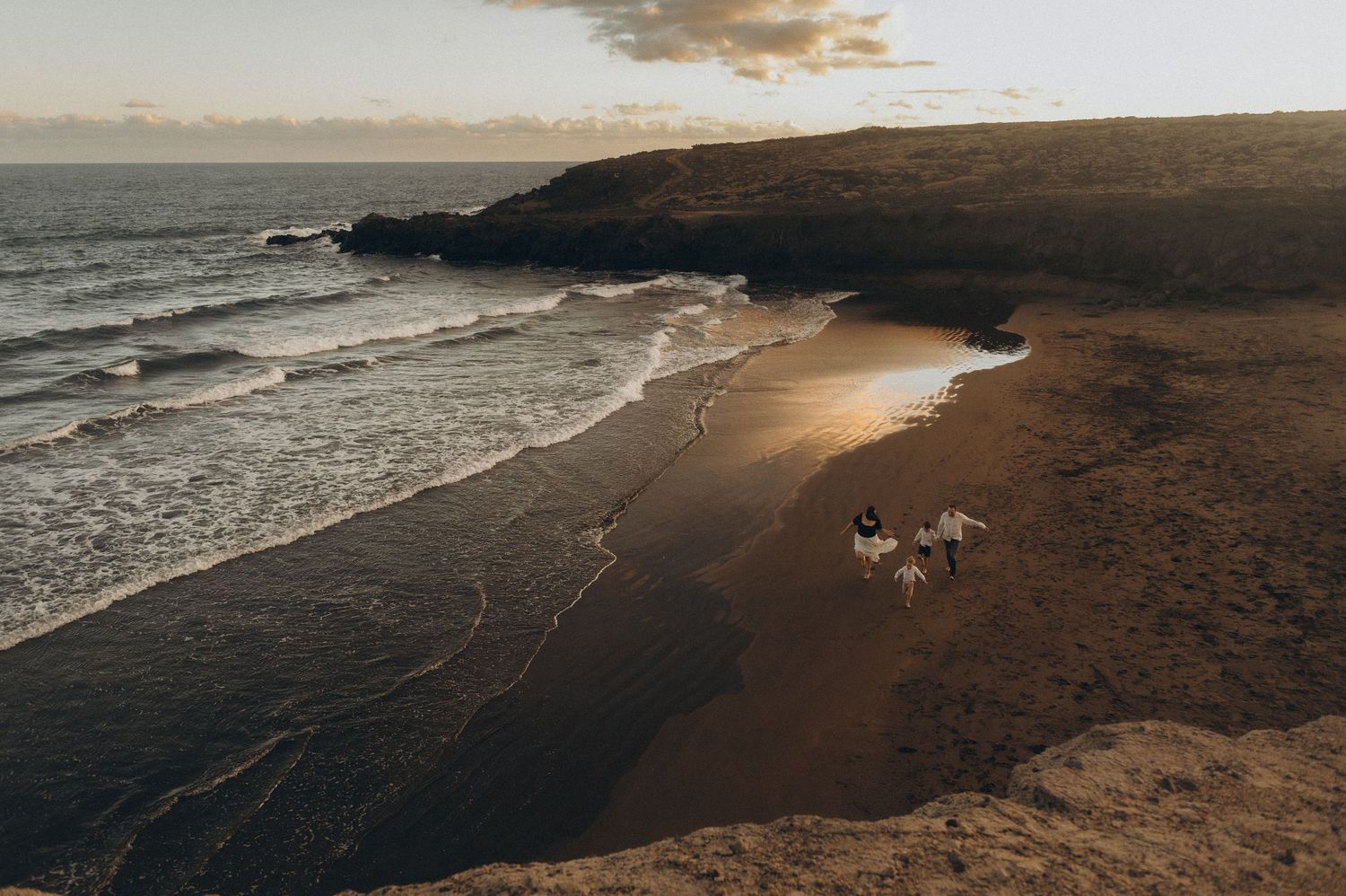 Aerial view of a couple walking on a secluded sandy beach at sunset with dramatic cliffs and ocean waves.