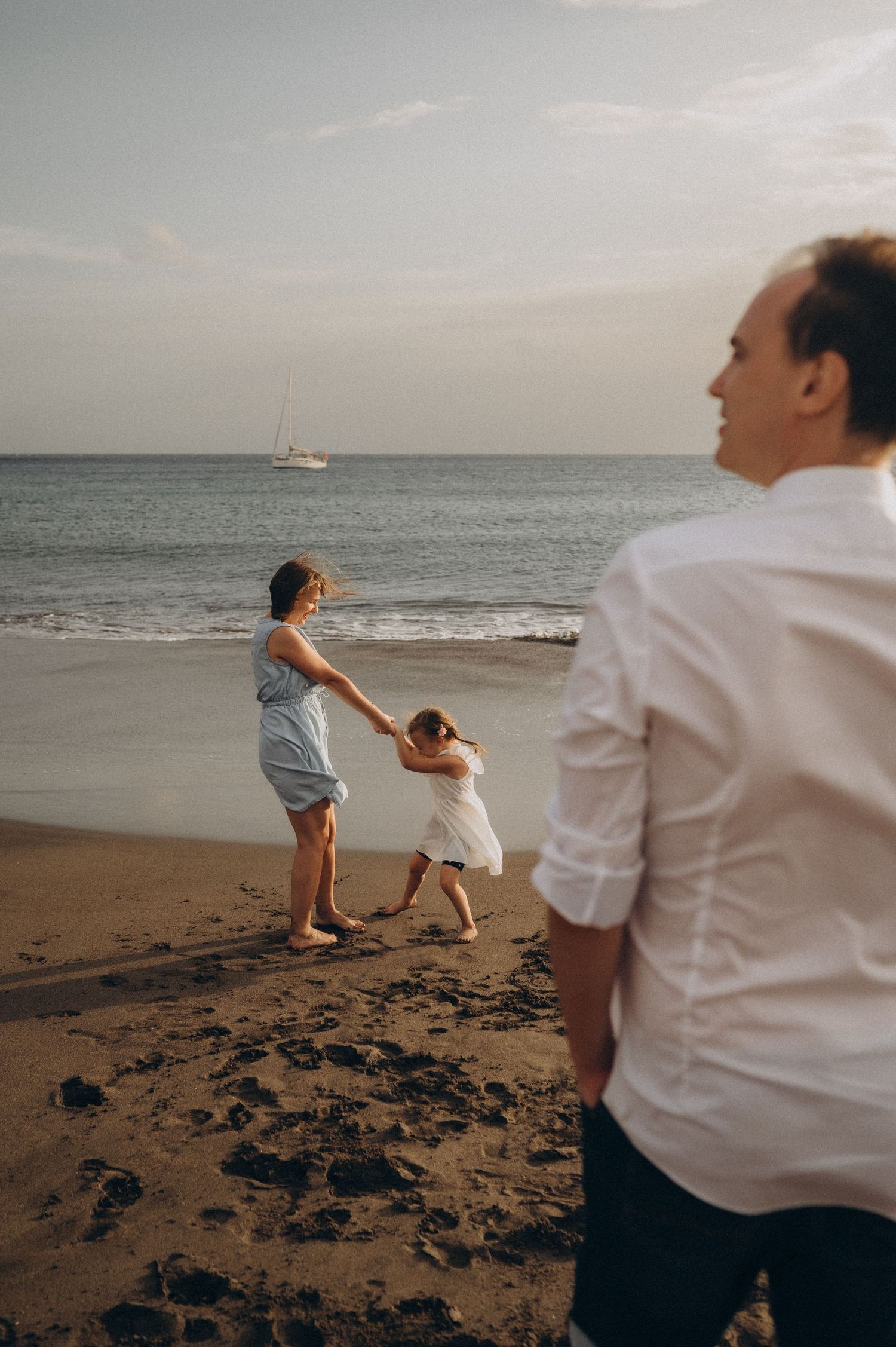 People throwing rocks into the ocean at sunset on a beach with a sailboat visible in the distance.