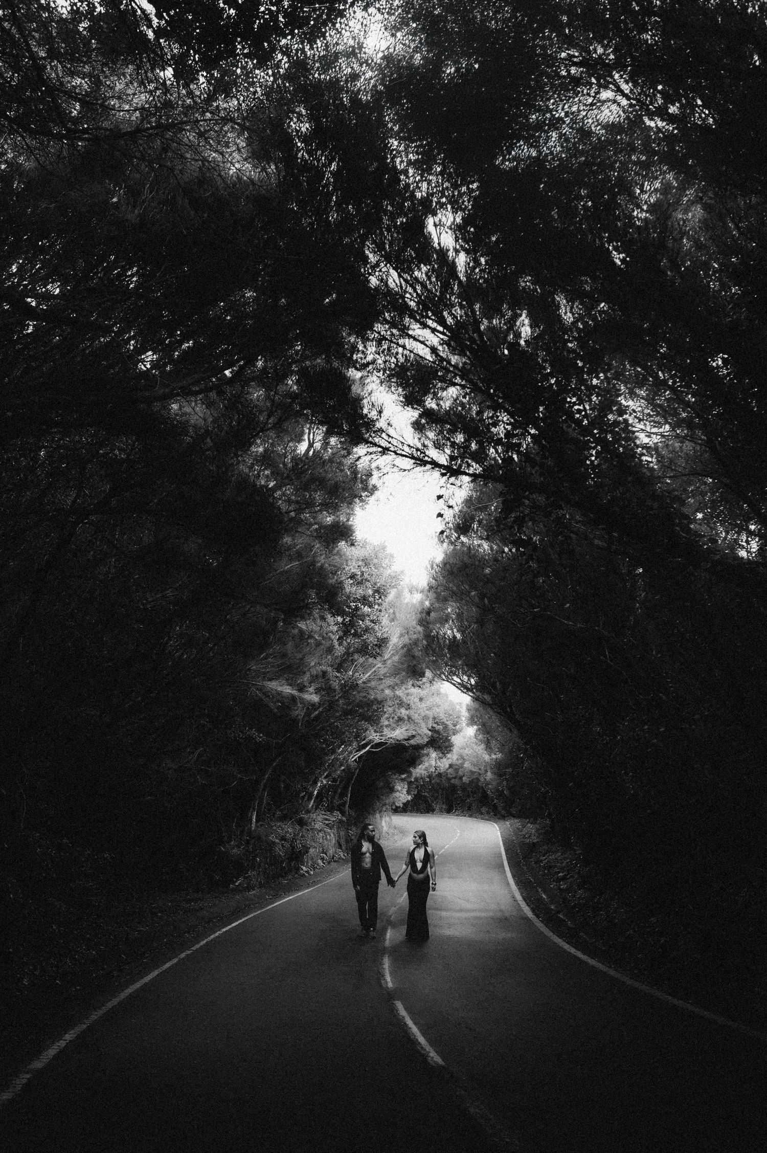 A couple holds hands walking down a tree-lined road in a romantic black and white forest photograph.