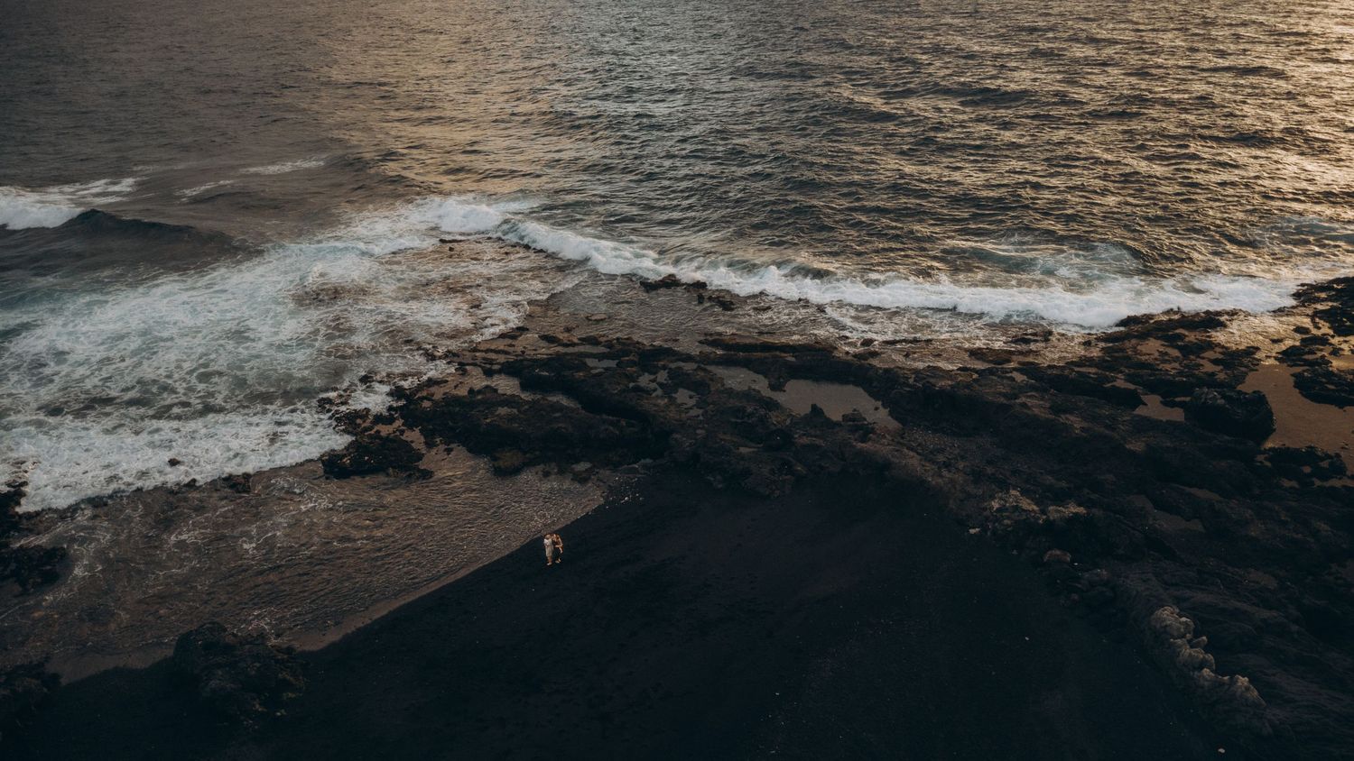 Aerial view of ocean waves crashing on rocky coastline at sunset with a lone figure standing on the shore.