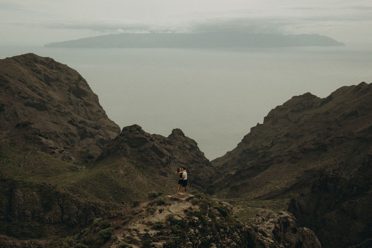 Two people stand on a rocky mountain ridge with misty ocean and island views in the background.