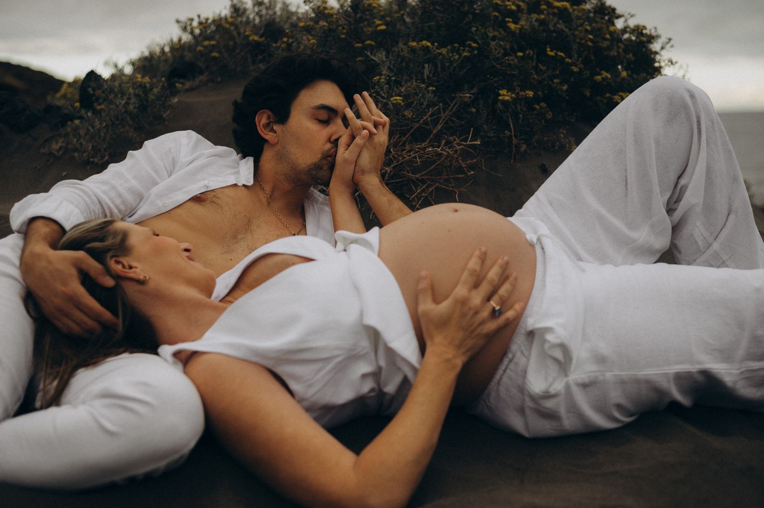Romantic couple in white outfits sharing a tender kiss outdoors, woman with baby bump lying together on the ground.