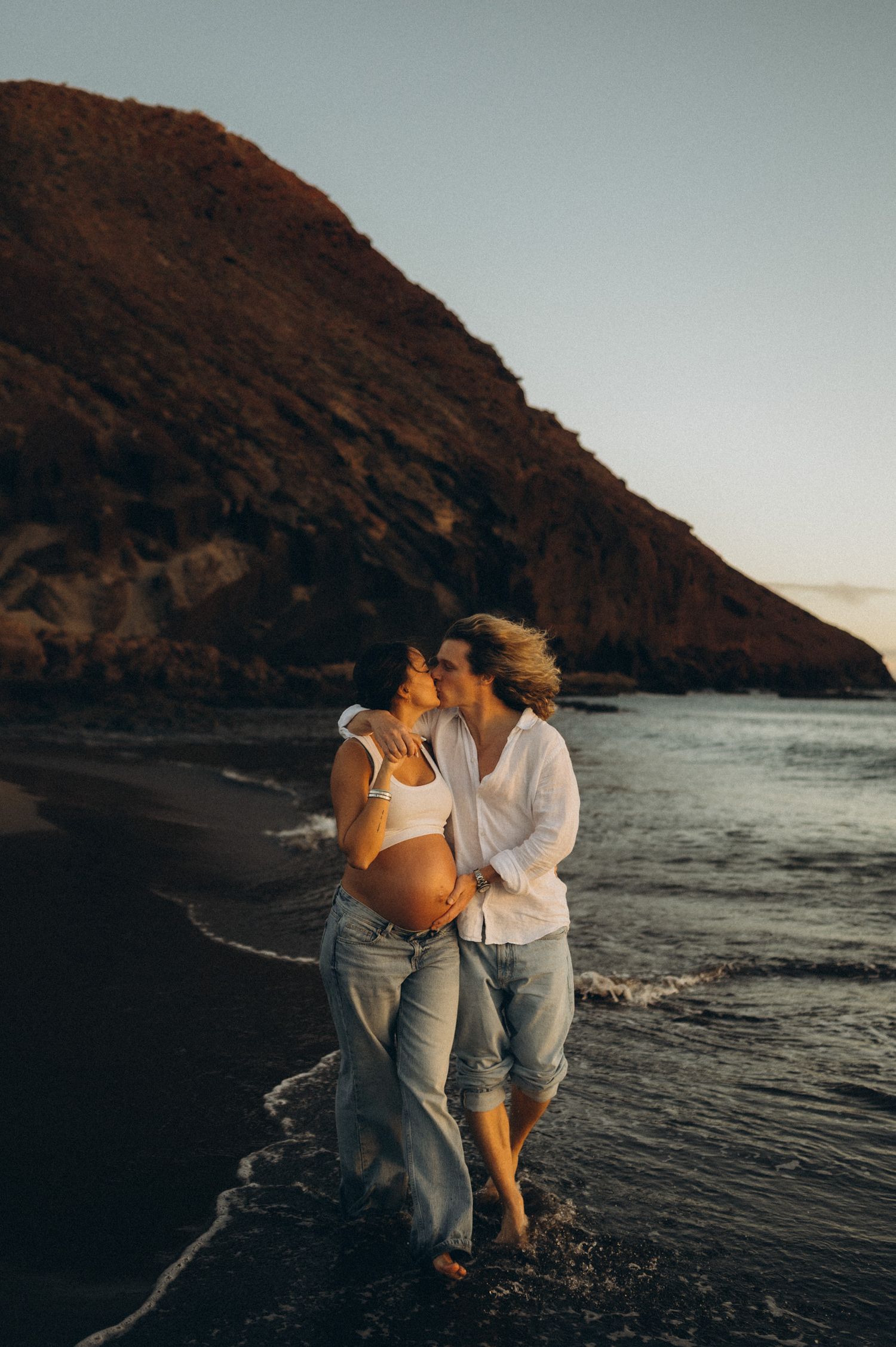 Pregnant couple kissing on a black sand beach at sunset with dramatic cliffs in the background.