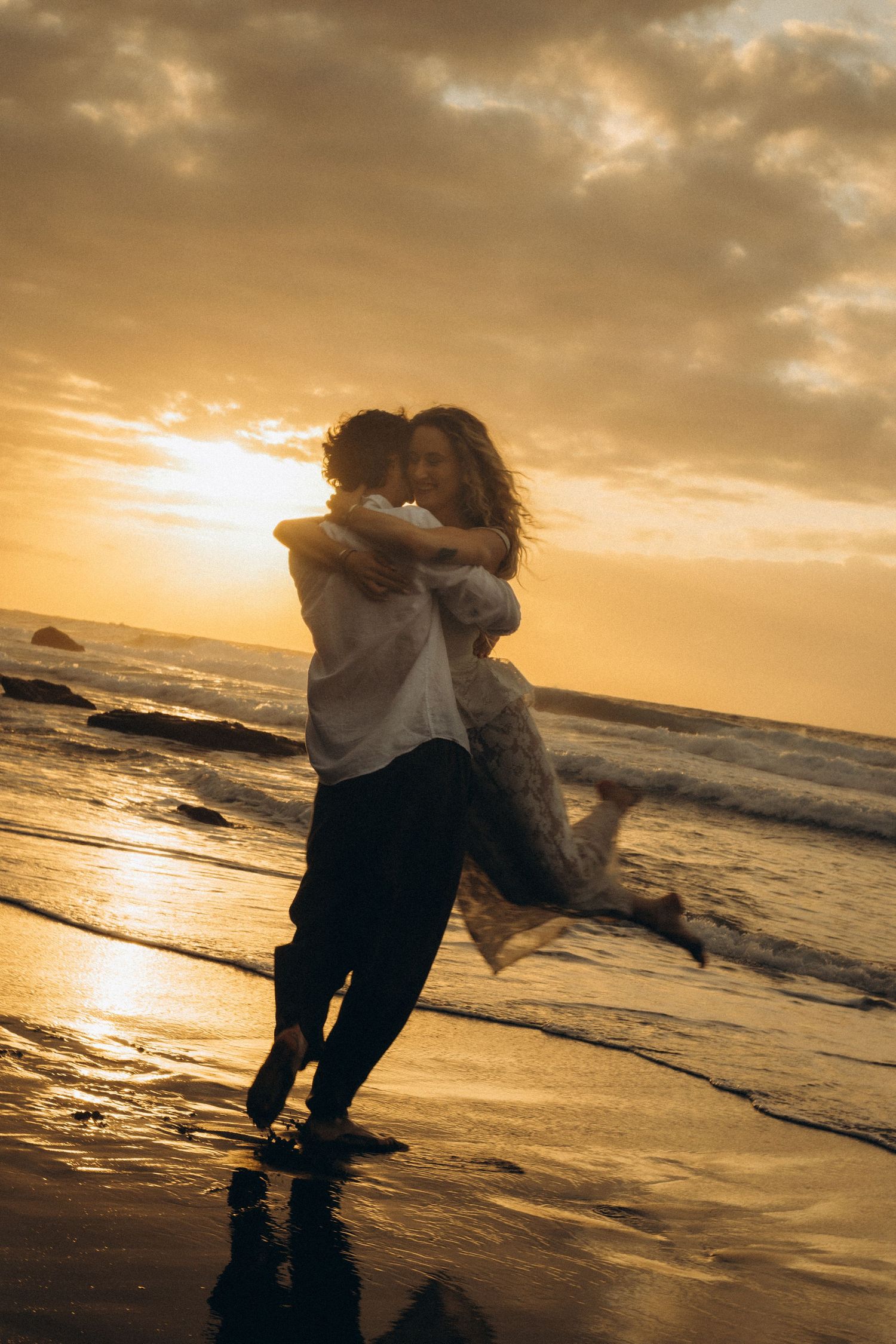 Couple embracing on a beach at golden sunset, waves reflecting warm light as man lifts woman in romantic seaside moment.
