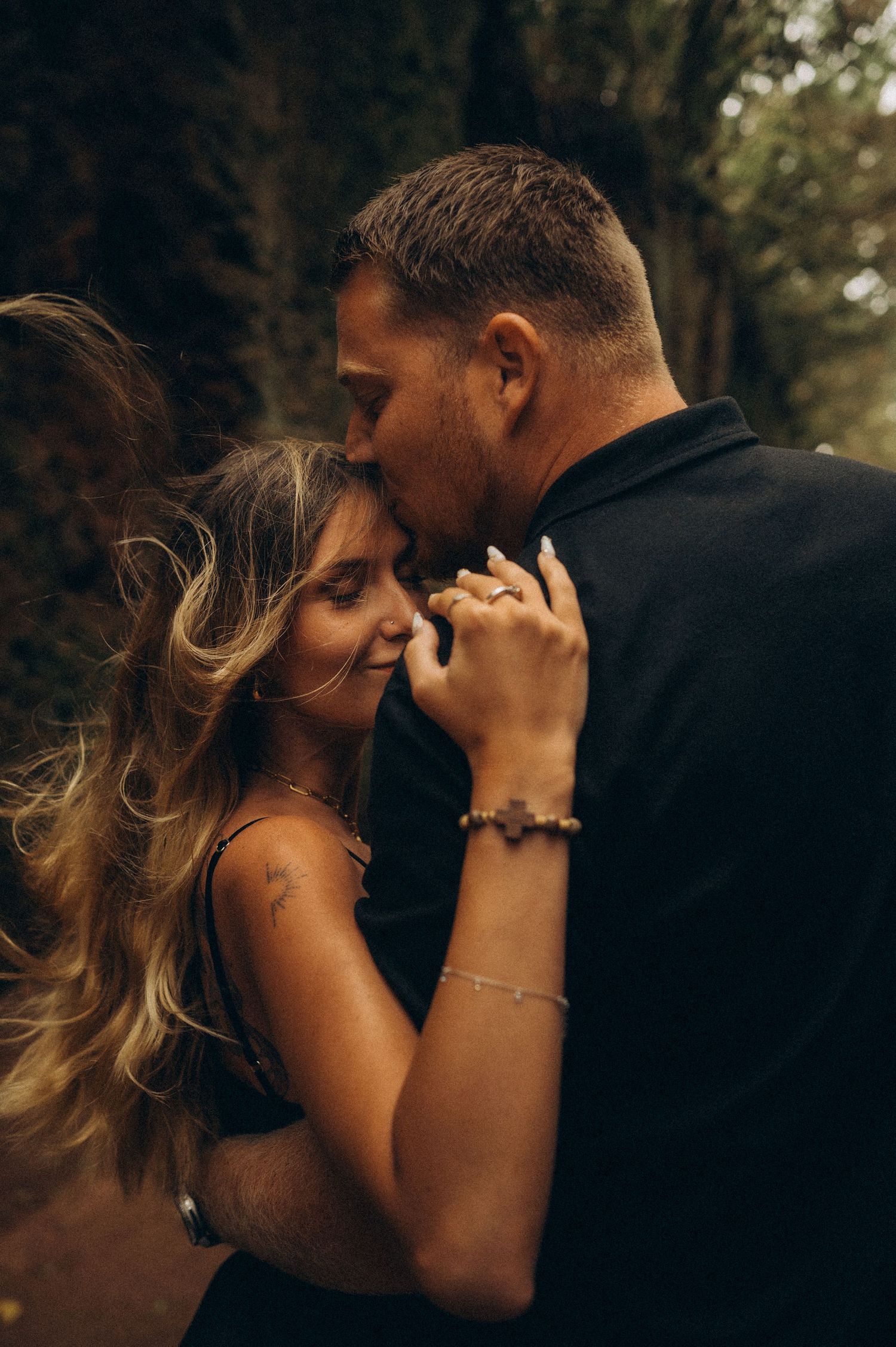 A romantic couple in black outfits share an intimate moment, holding hands and touching foreheads outdoors.