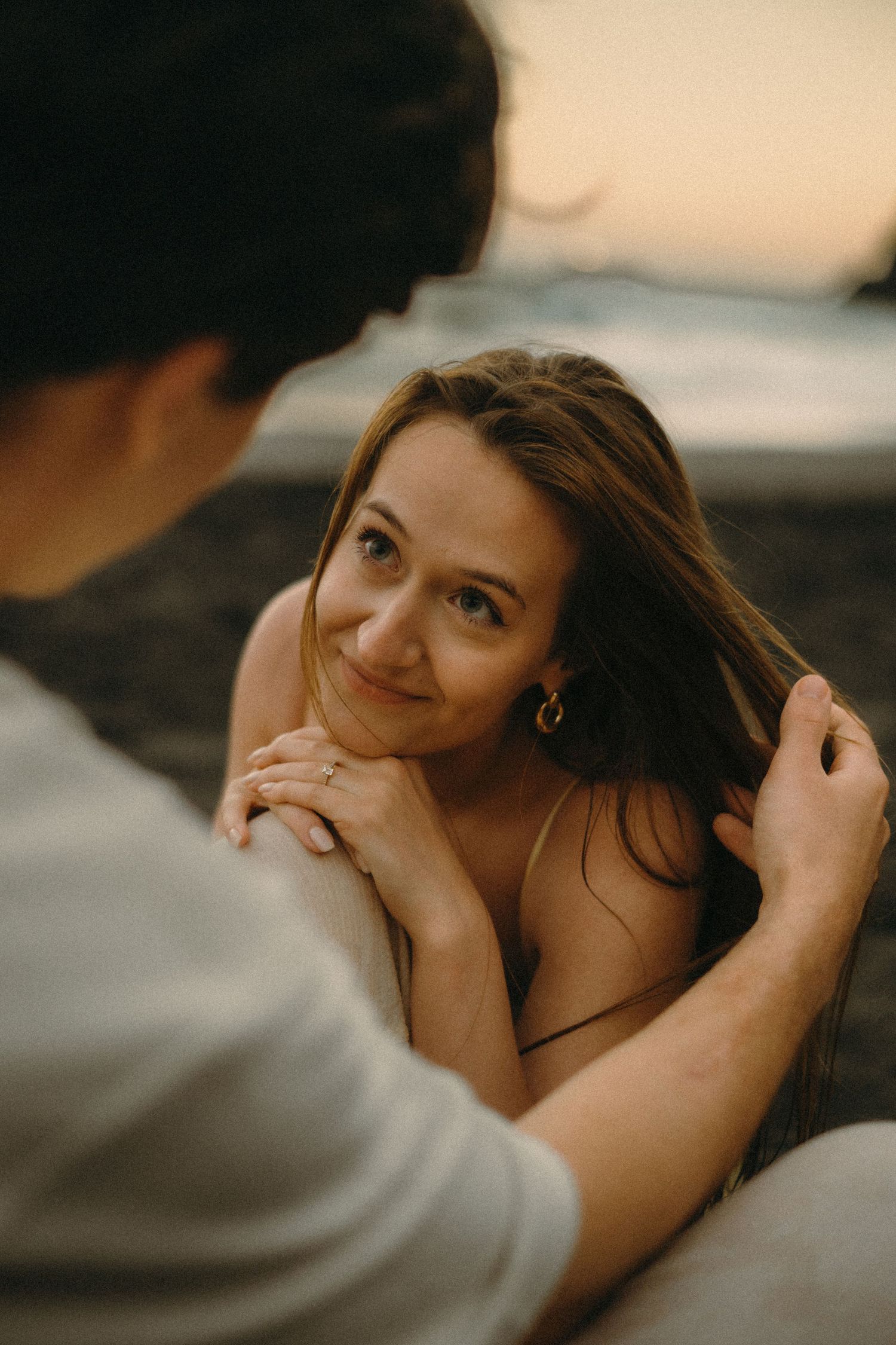 A woman with long brown hair smiles warmly at a man on a beach at sunset, resting her chin on her hands.