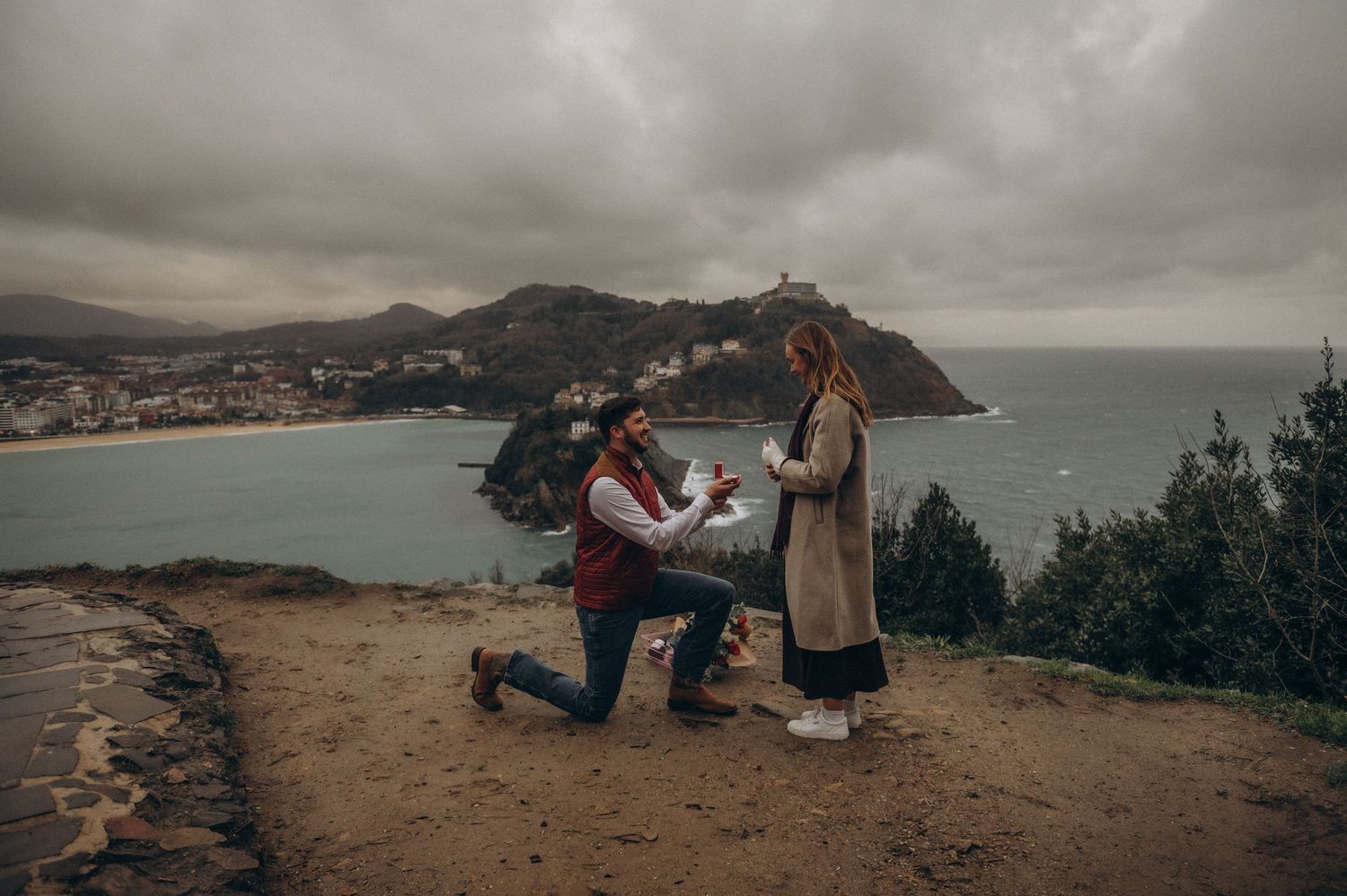 Surprise proposal and engagement in Tenerife on a scenic coastal overlook with a bay and hills in the background.