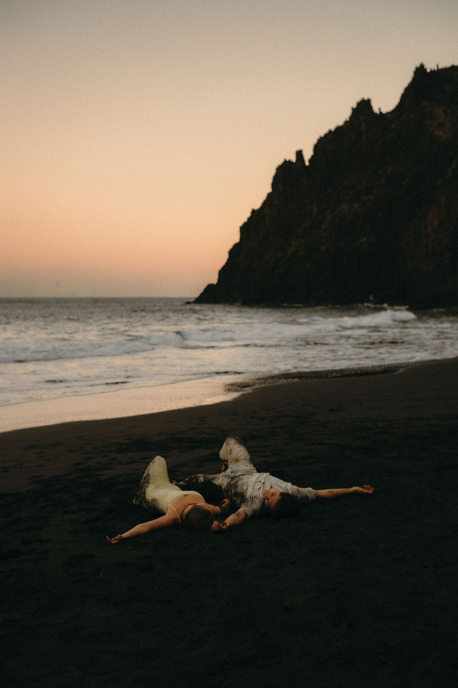 Couple lying on black sand beach at sunset with dramatic rocky cliffs and ocean waves in background.