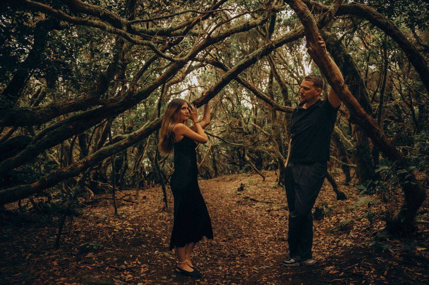 Couple sharing a romantic moment in a moody forest, surrounded by twisted branches and autumn leaves.