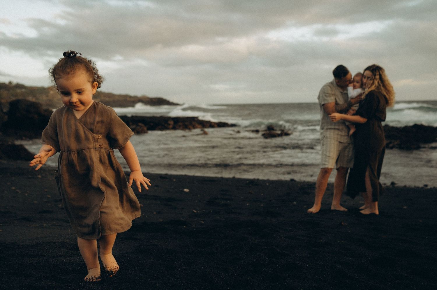 Young girl plays on black sand beach while parents embrace near ocean waves in moody coastal family photo.