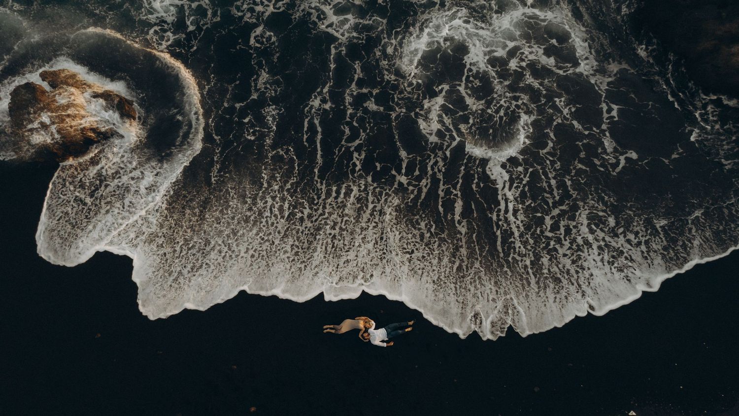 Aerial view of dark ocean waves crashing against rocky coastline creating dramatic patterns of white foam.