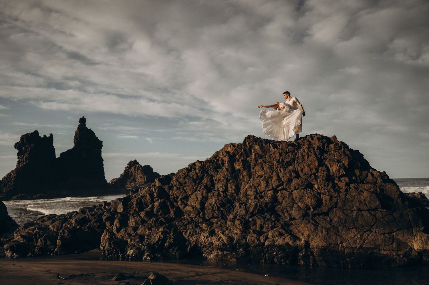 Distant shot of couple on large volcanic rock with cliffs and Atlantic Ocean at Playa Benijo, Tenerife