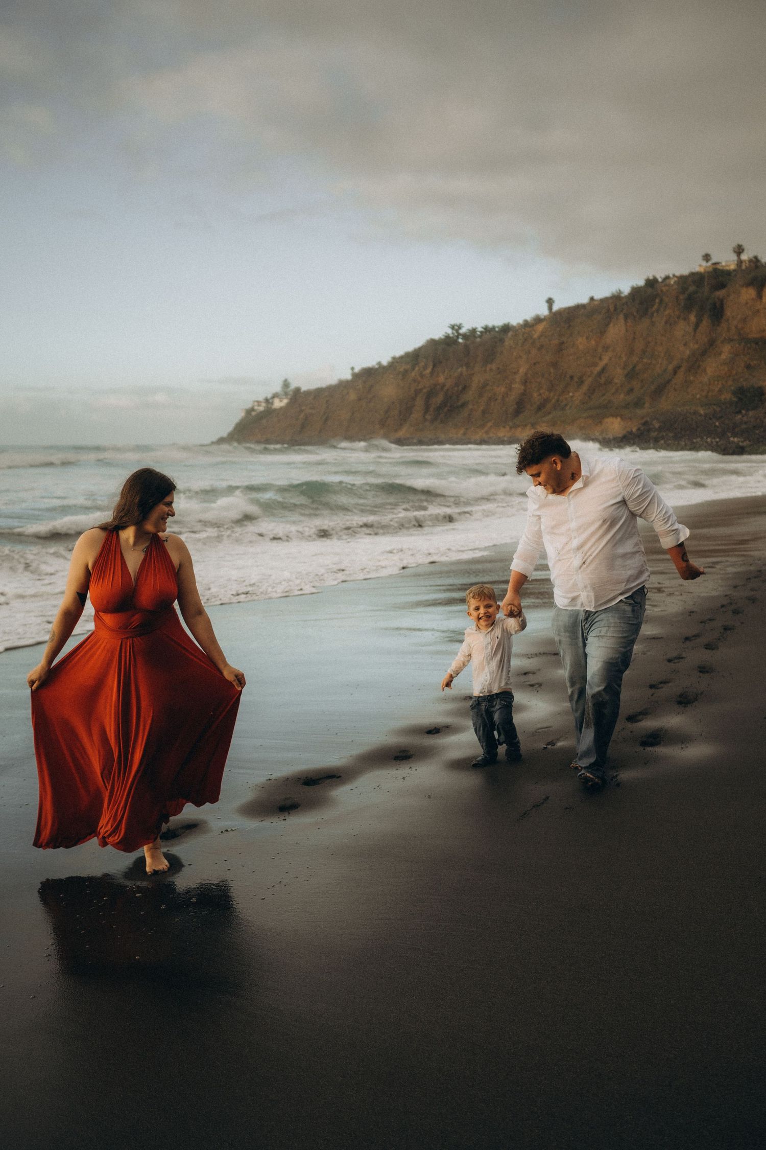 Family of three walking on a sandy beach at sunset, woman in flowing red dress, man in white shirt, with young toddler.