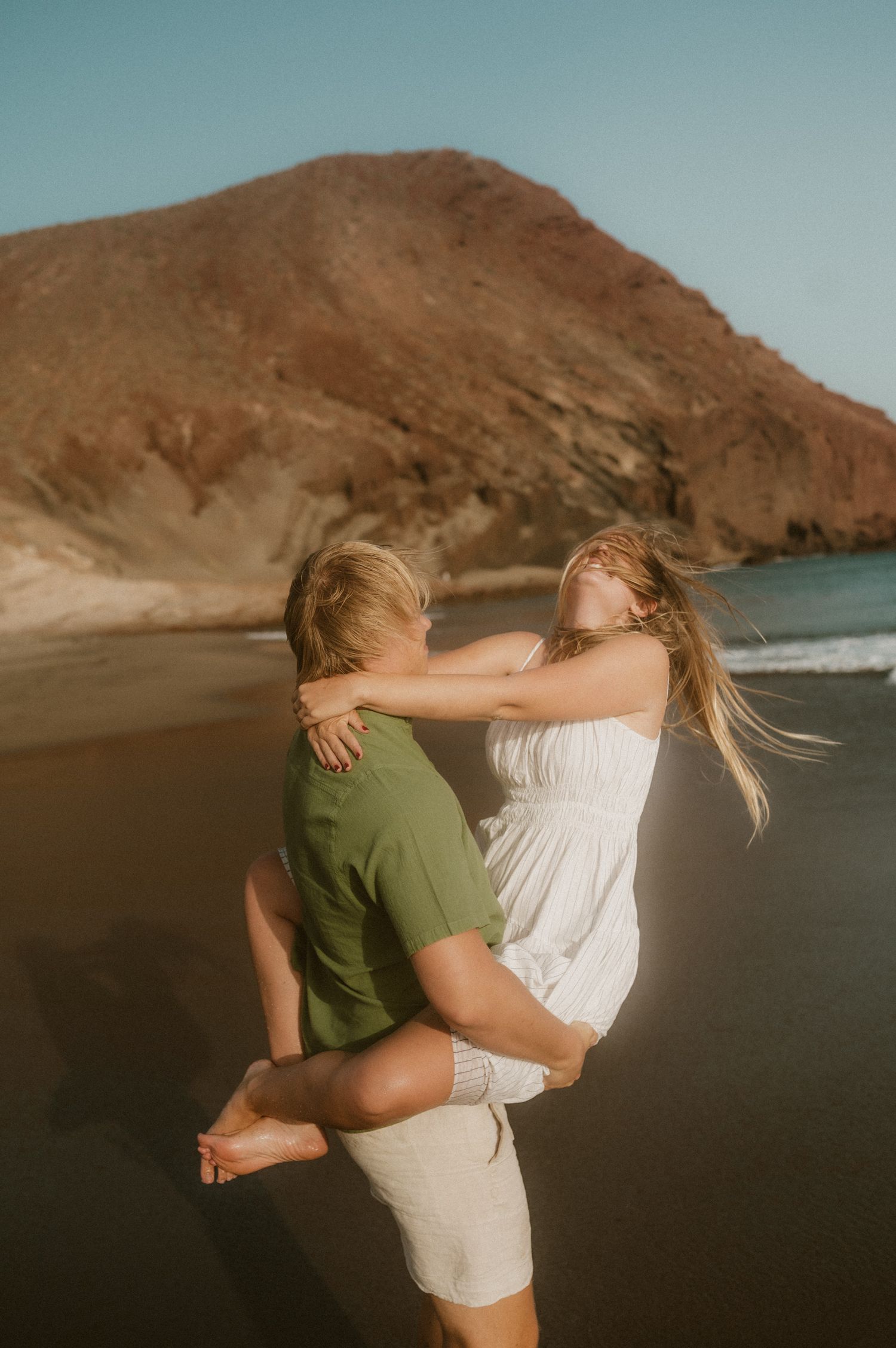 Young couple on Playa Tejita, he holds her firmly in his arms while she leans back enjoying the moment, her hair flowing in the wind, Montañ