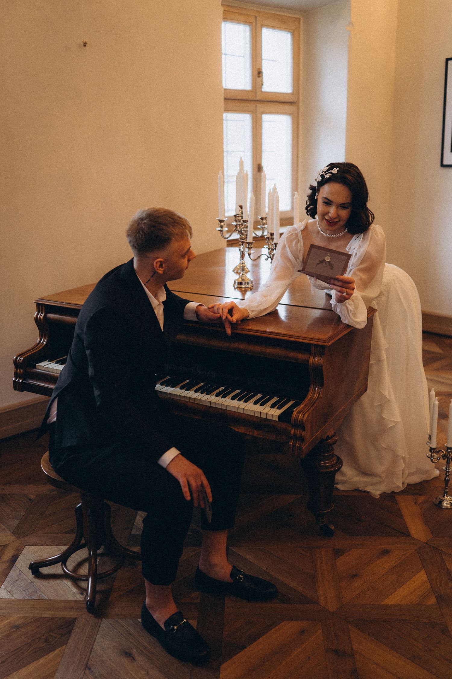 Romantic couple portrait with the groom seated at a piano and the bride leaning elegantly against it as they look at each other.