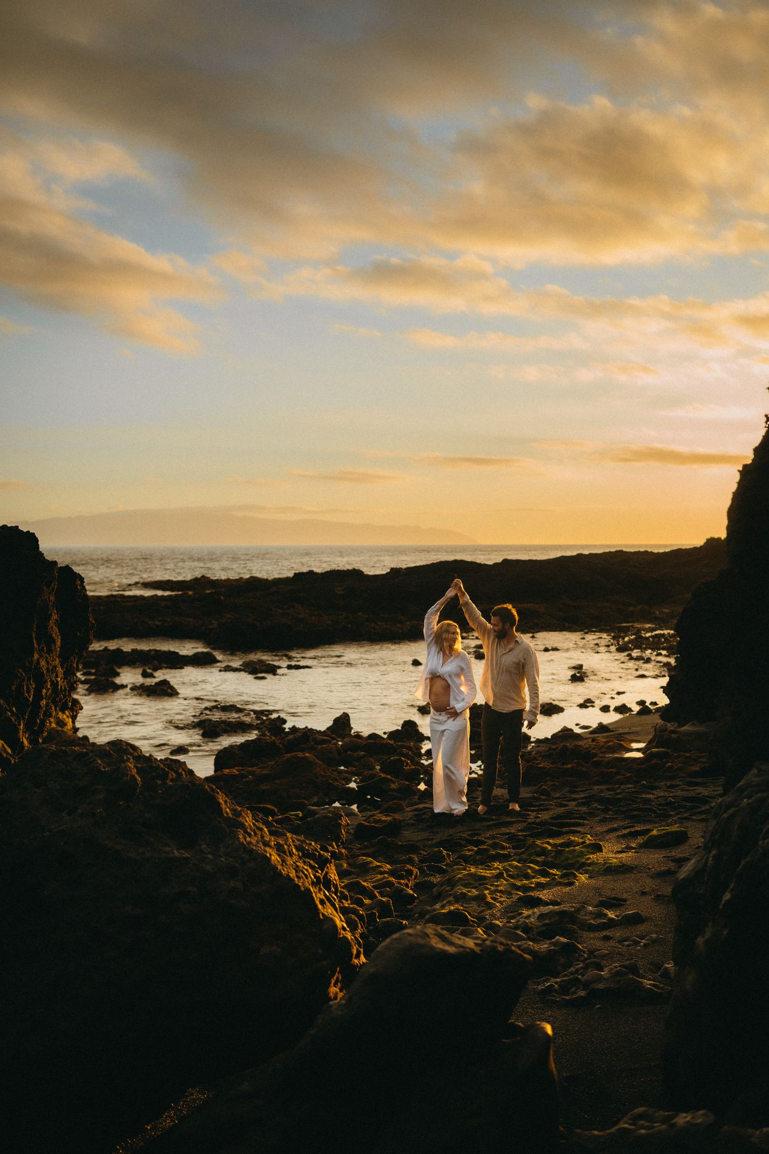 Couple dancing on rocky beach at golden sunset with ocean waves and dramatic cloudy sky in background.