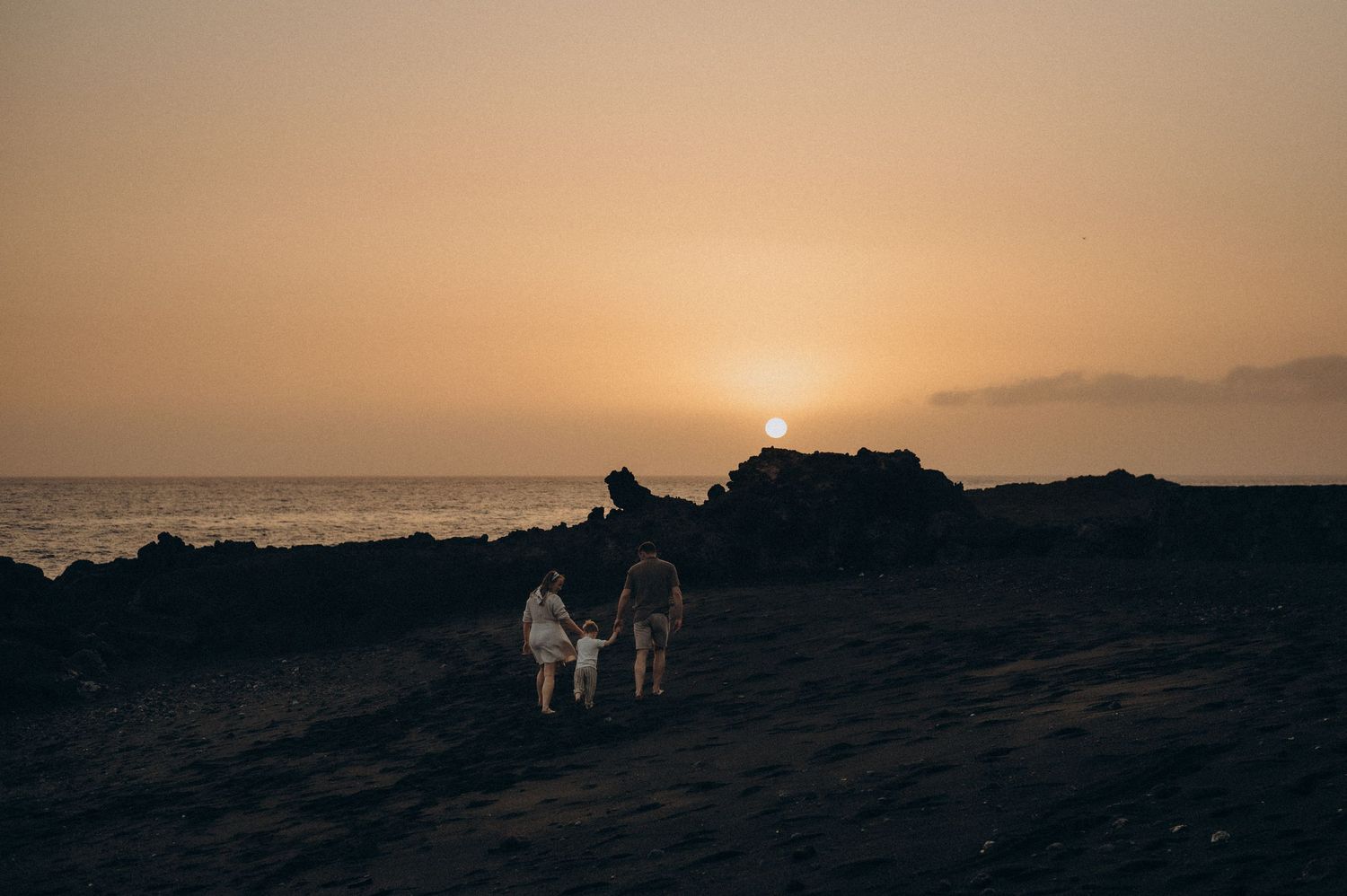 Family silhouetted against a golden sunset on a rocky beach, with calm ocean waters in the background.