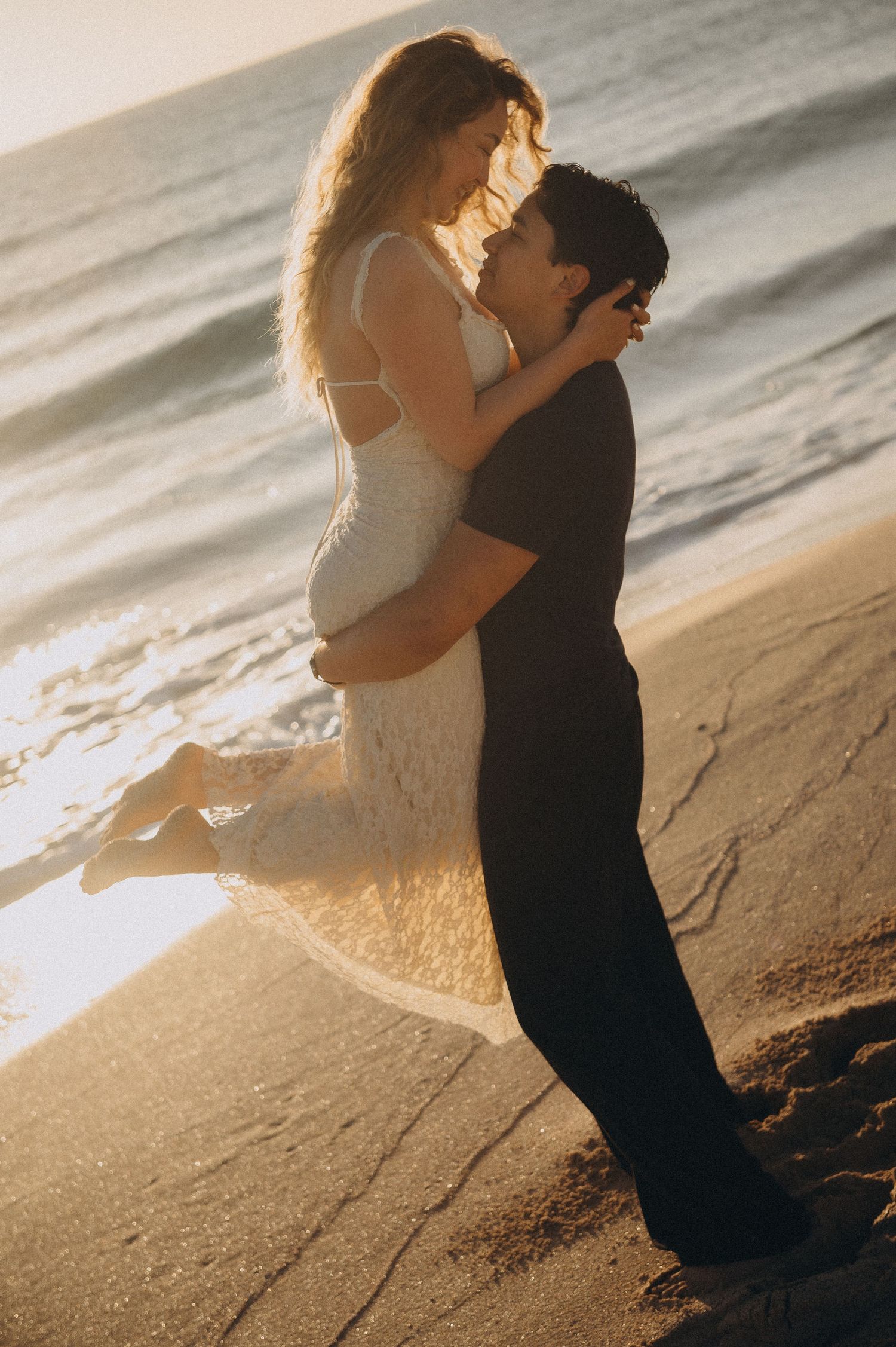 A couple embraces romantically on a sandy beach at golden sunset, the man lifting the woman in a white dress by the ocean.