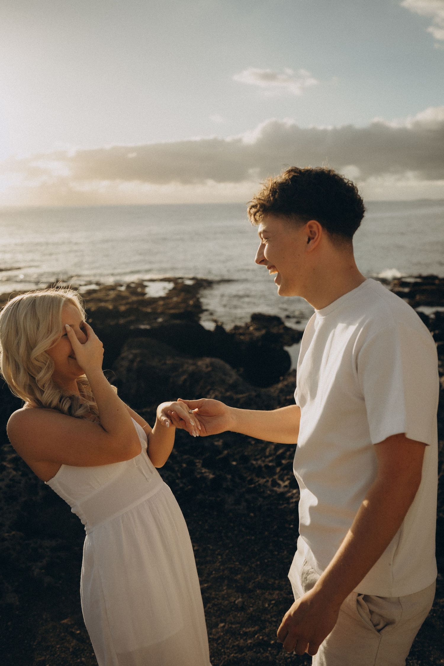 Couple holding hands at sunset on rocky ocean shore, woman in white dress, man in white shirt, romantic coastal scene.