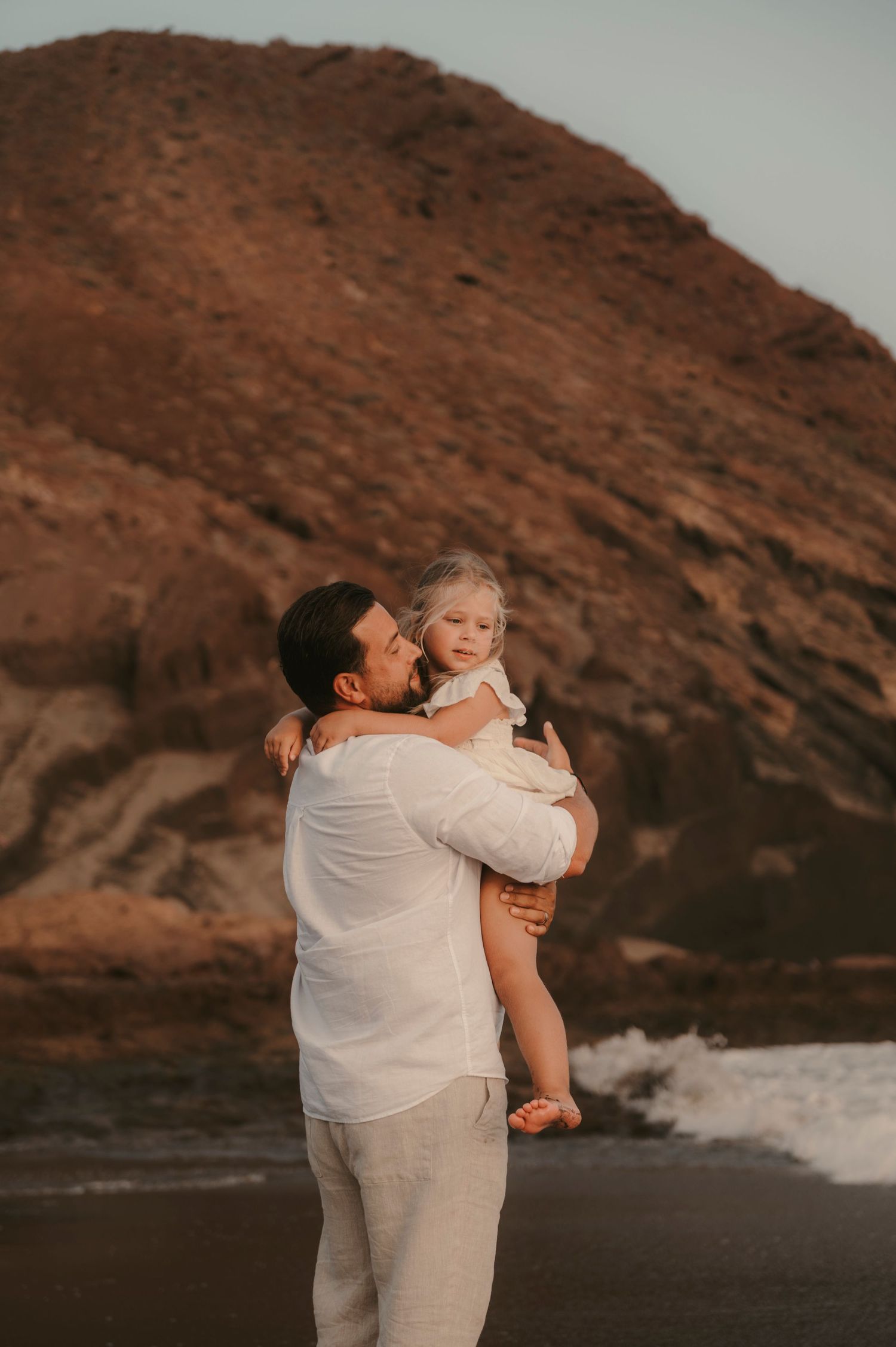 Father holding his daughter in his arms with majestic Montaña Roja in the background at Playa La Tejita