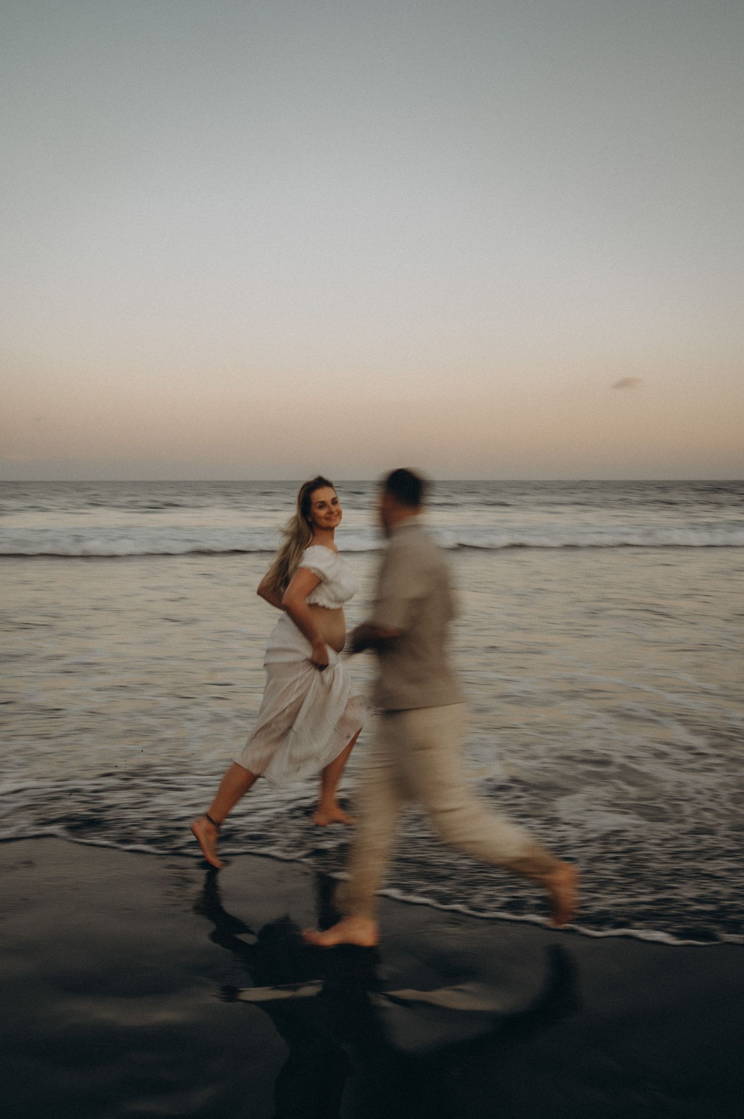 A couple runs barefoot on a dark sandy beach at sunset, the woman in white smiling joyfully as waves roll in behind them.