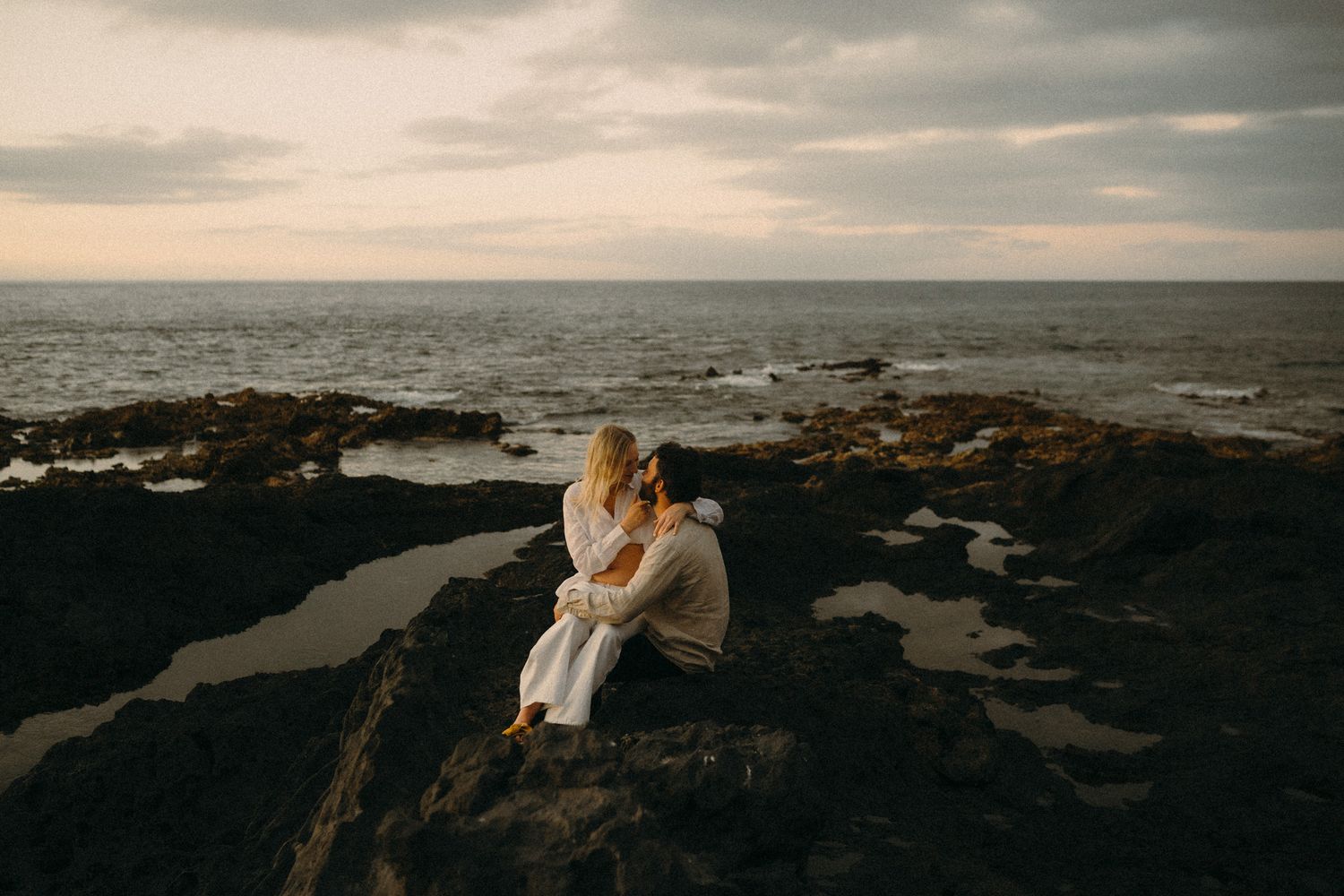 Couple embracing on rocky coastal cliffs at sunset with dramatic ocean waves and cloudy sky in background.