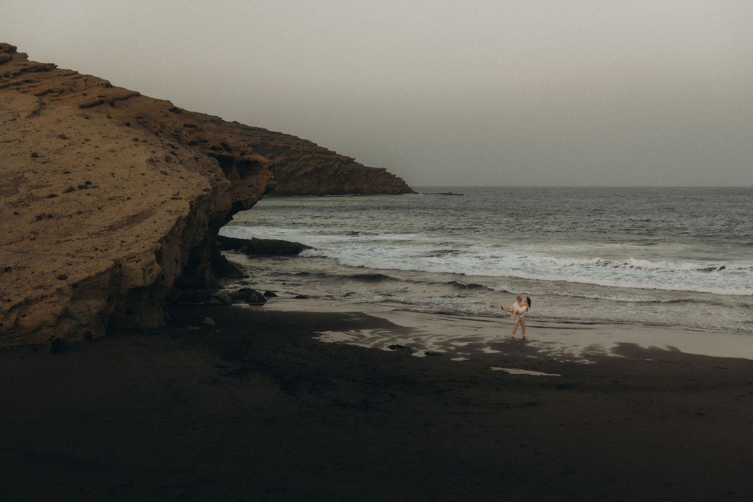 A lone figure stands on a dark volcanic beach beside dramatic cliffs with ocean waves under a moody overcast sky.