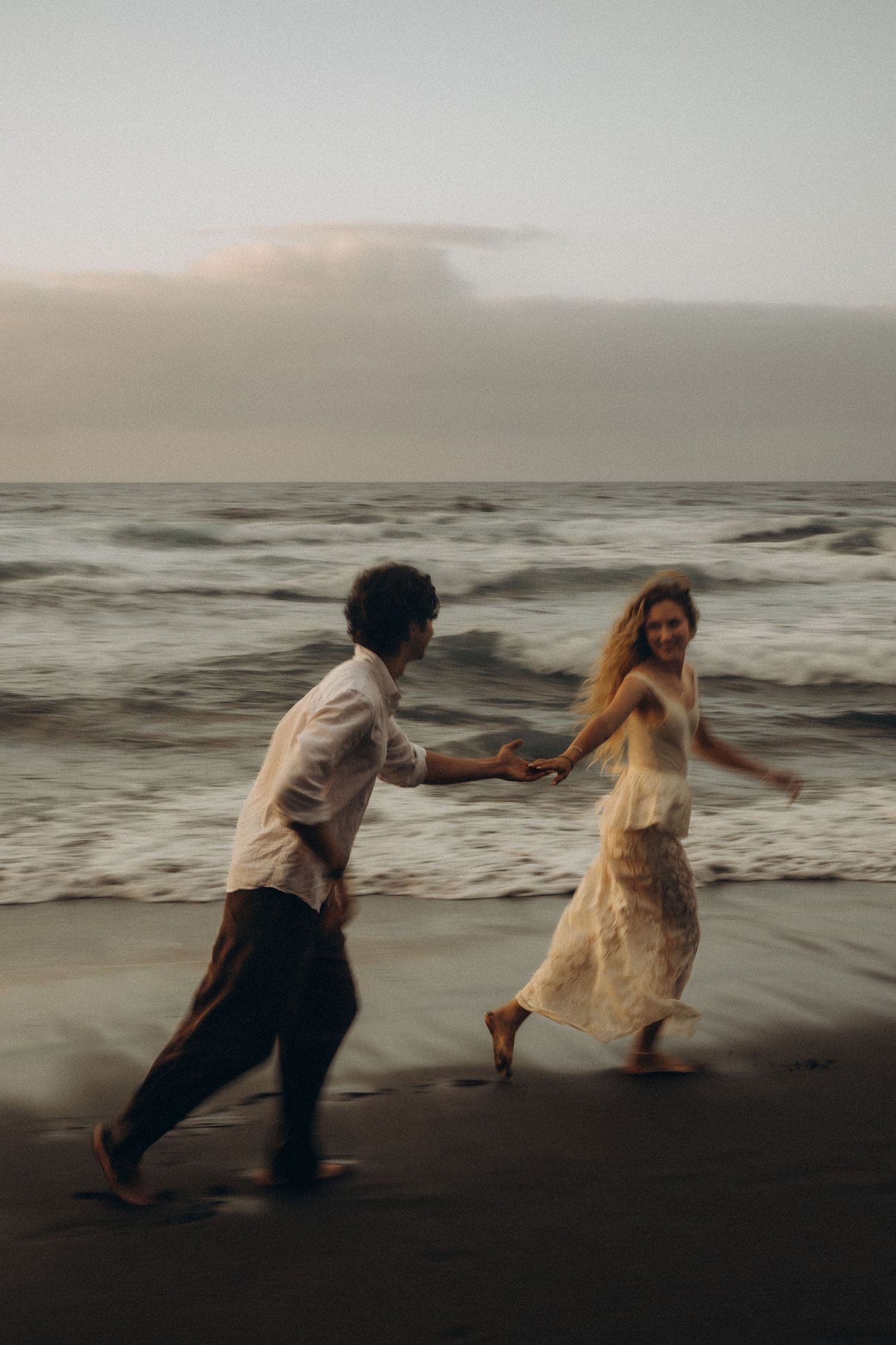 Couple holding hands running barefoot on a dark sandy beach at dusk with ocean waves in the background.
