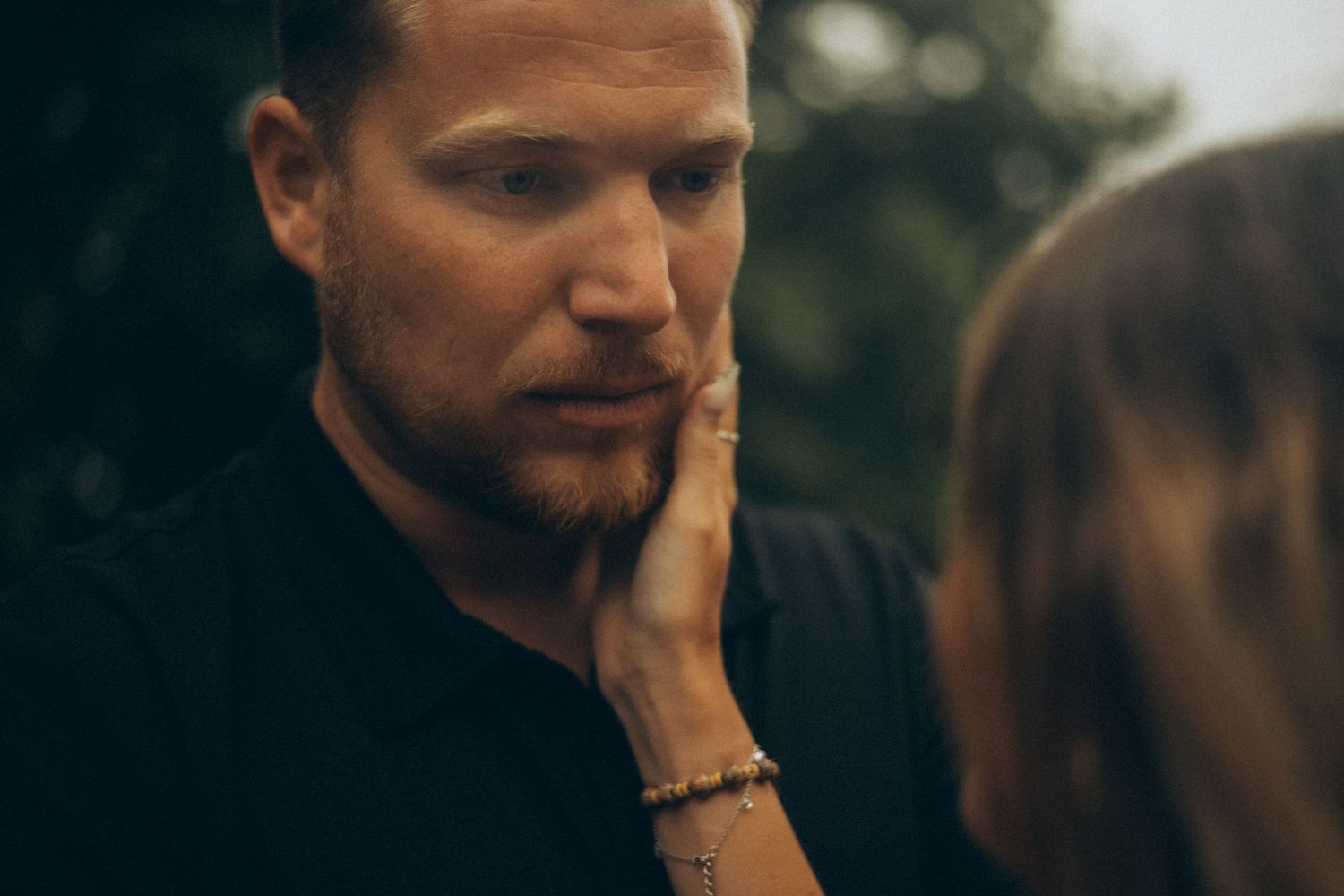 A woman gently touches a bearded man's face outdoors in an intimate moment.
