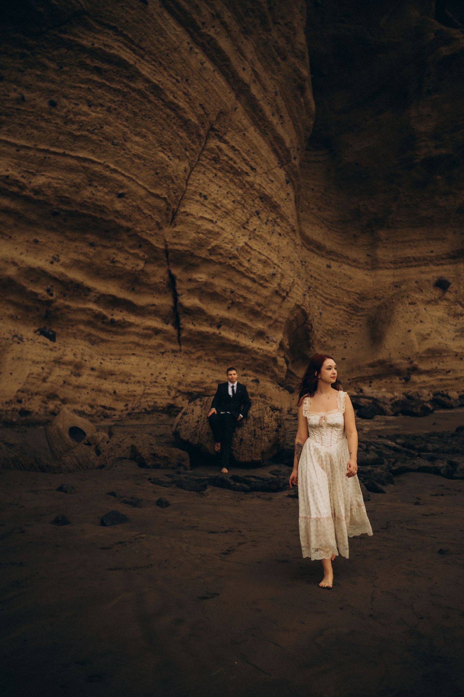Bride in white dress walks barefoot through a dramatic sandstone cave while groom follows in dark suit.
