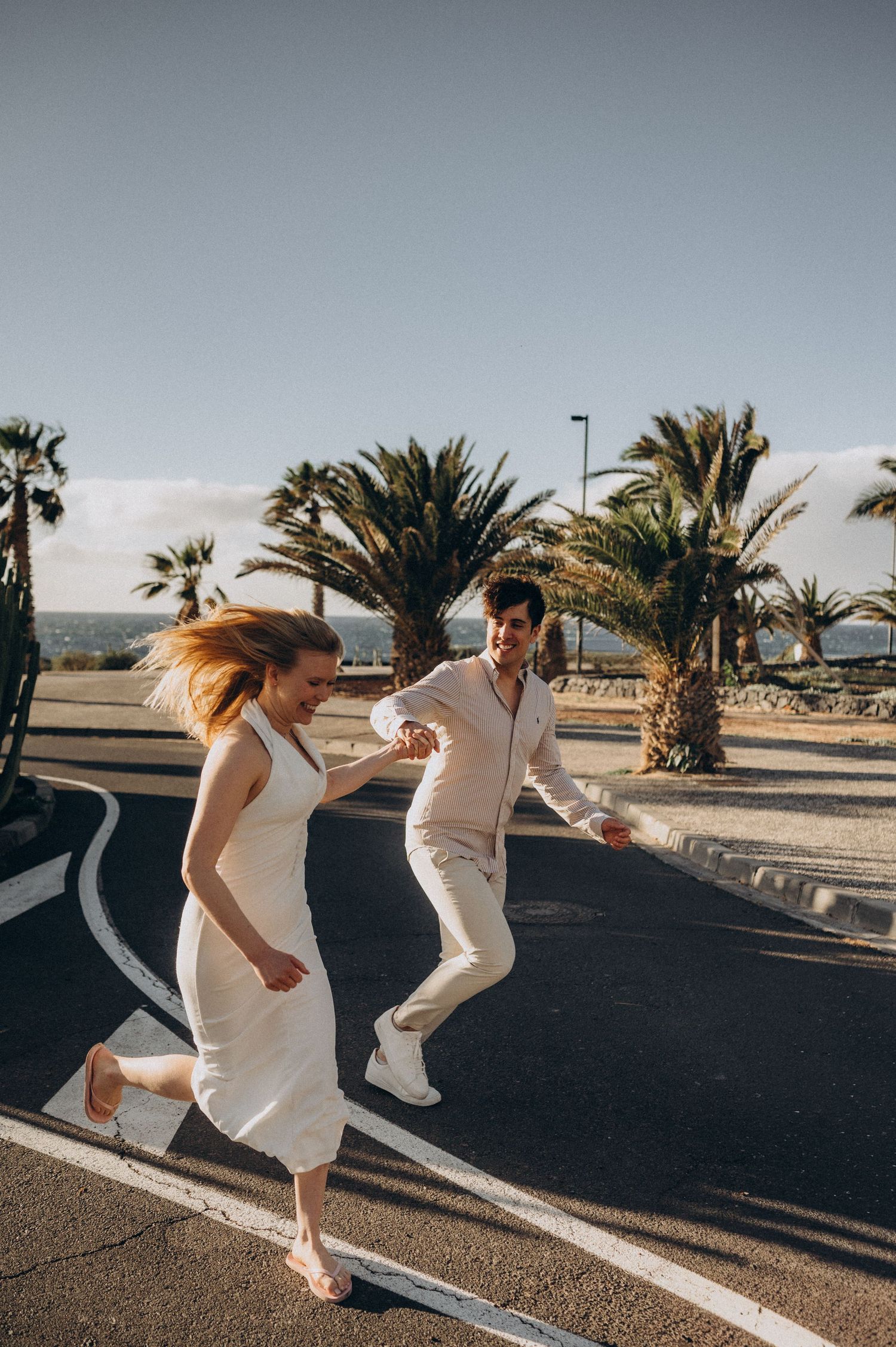 Couple in white outfits joyfully running hand-in-hand across a sunny parking lot with palm trees in the background.