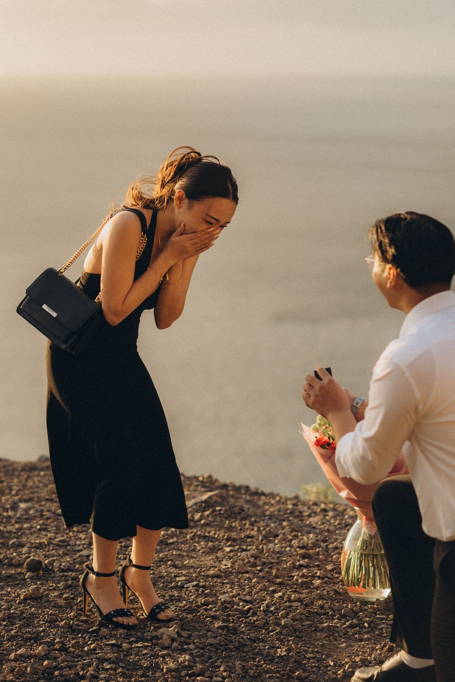 Man kneeling with flowers proposing to surprised woman in black dress on scenic coastal cliff at sunset.