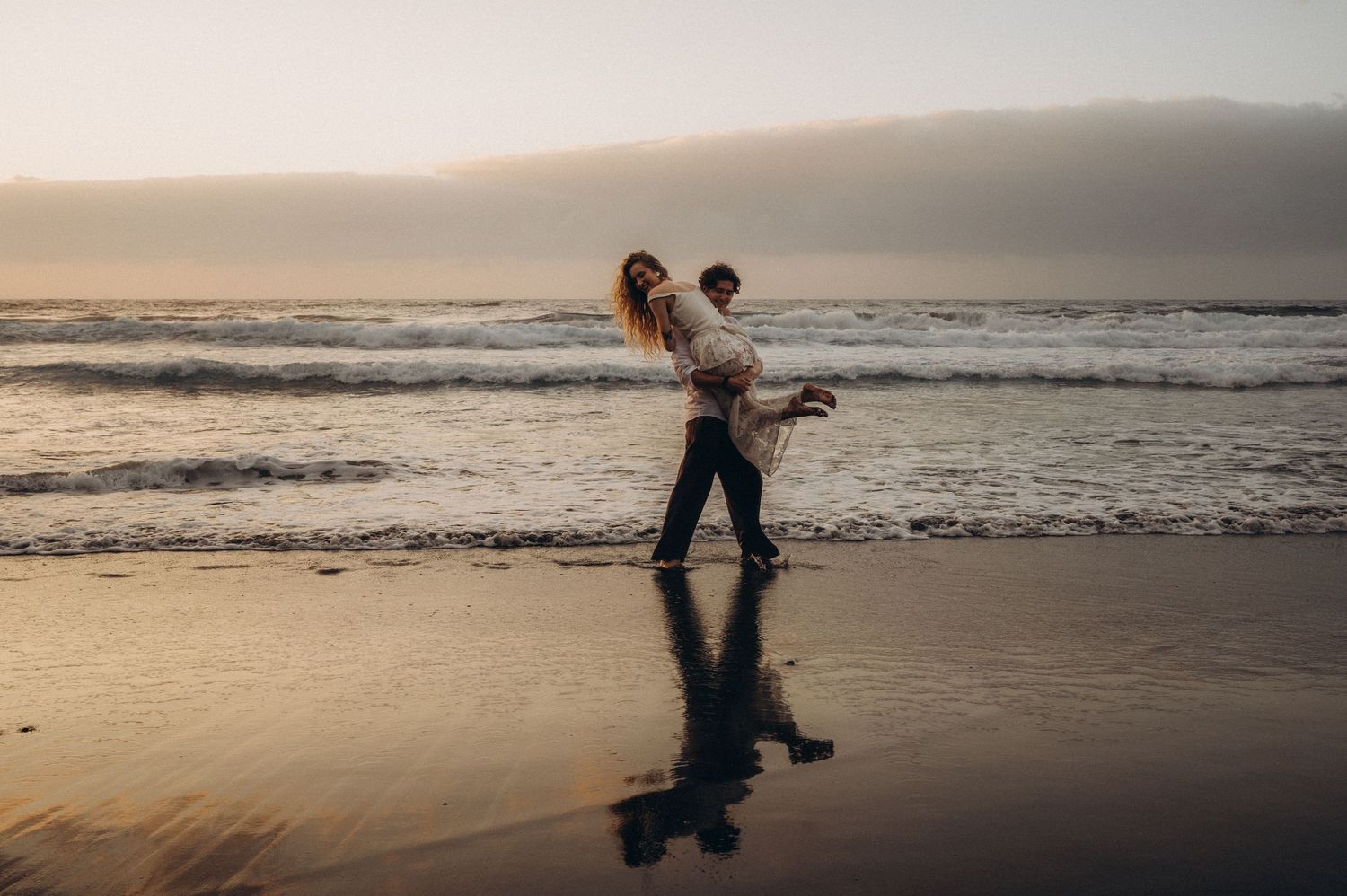 Couple sharing a romantic piggyback ride on a reflective beach at sunset with ocean waves in the background.