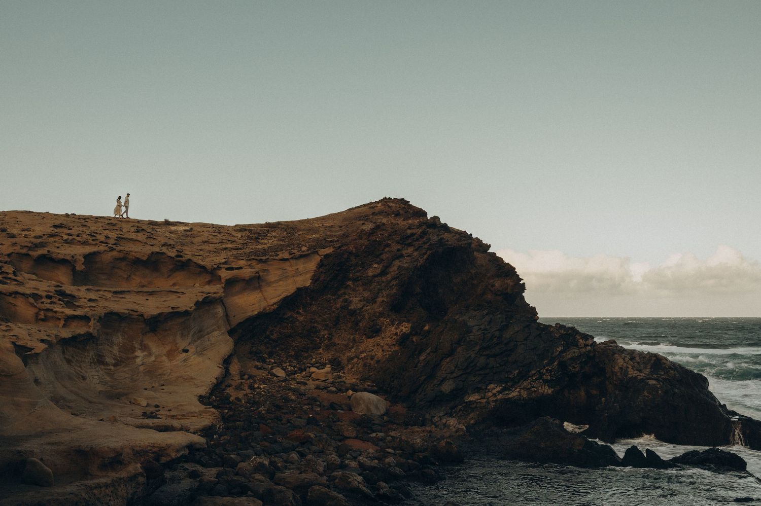 Two people stand atop a dramatic coastal cliff overlooking rocky shores and ocean waves at golden hour, Tenerife