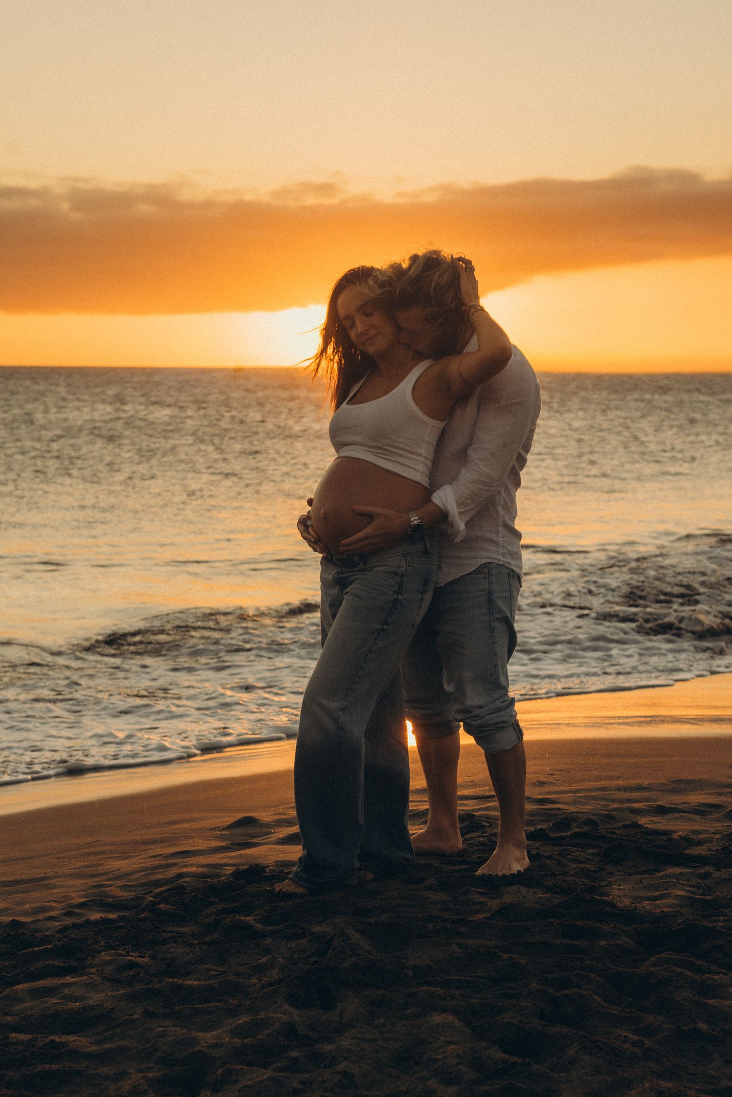 Couple embracing on a sandy beach at golden sunset, waves gently rolling in behind them.