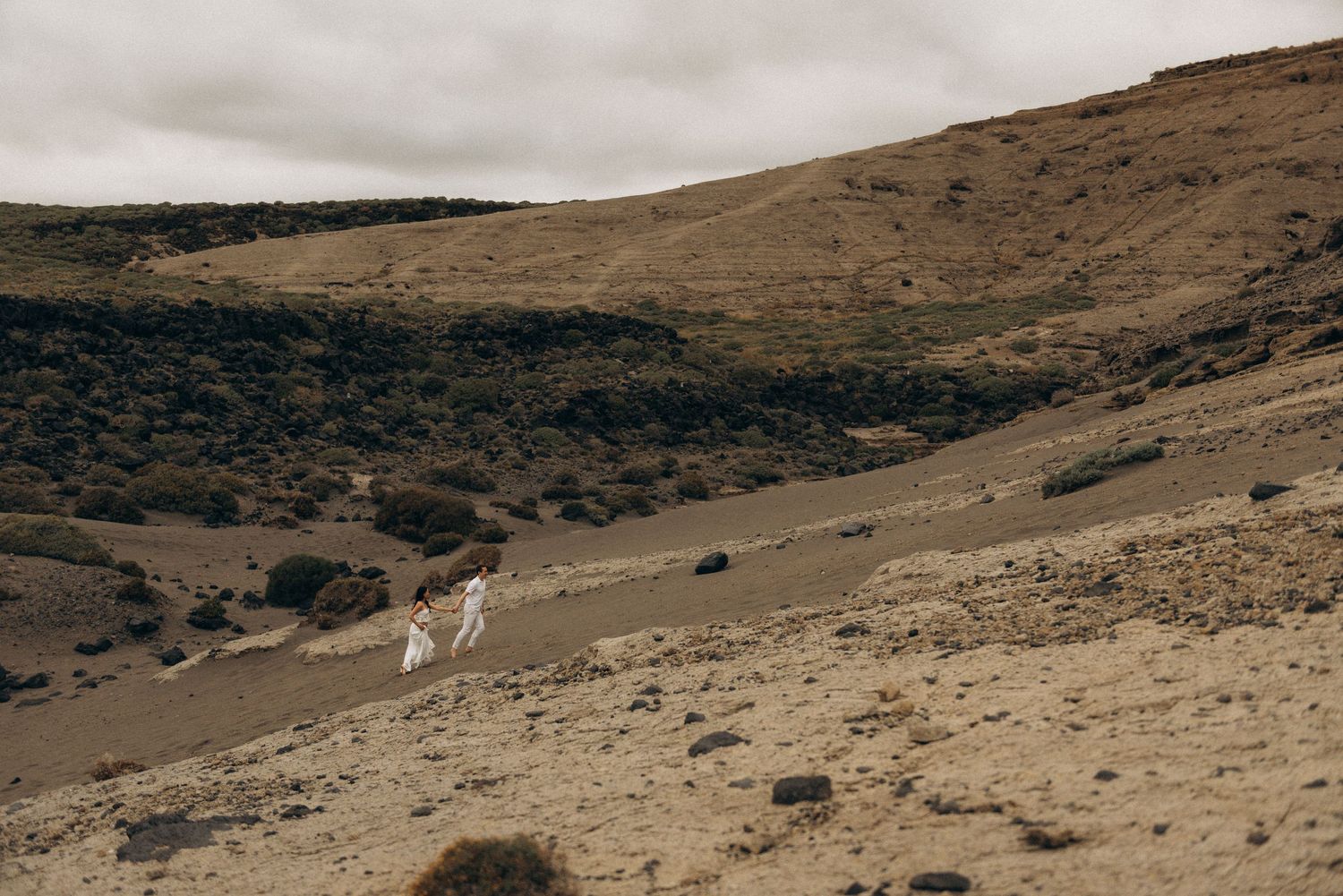 Two people in white attire walk along a sandy desert trail surrounded by arid hills under an overcast sky.
