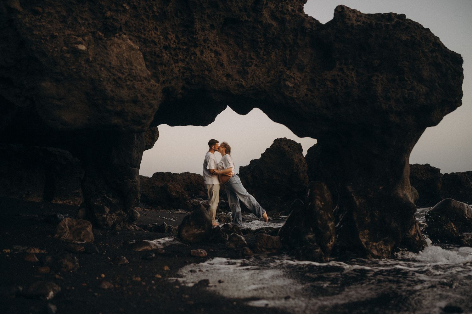 Couple embracing romantically under a natural rock arch on a rugged coastline at dusk.