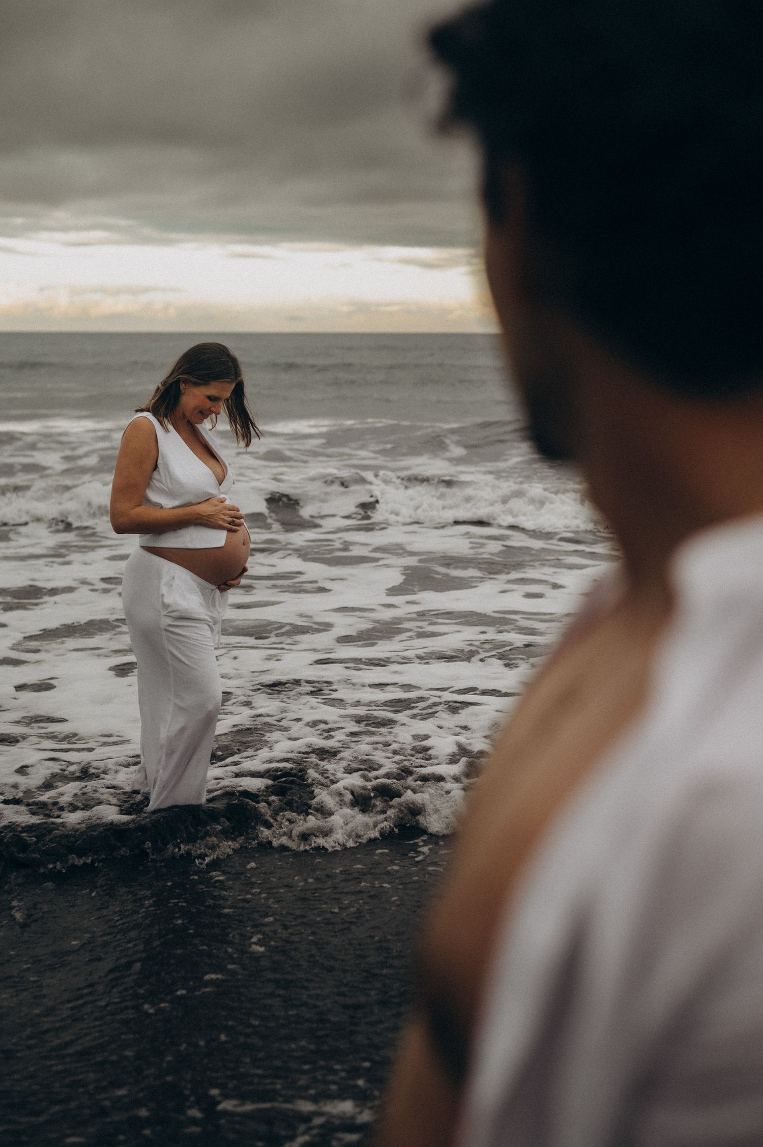 Pregnant woman in white dress standing in ocean waves at sunset, with a man in the foreground.