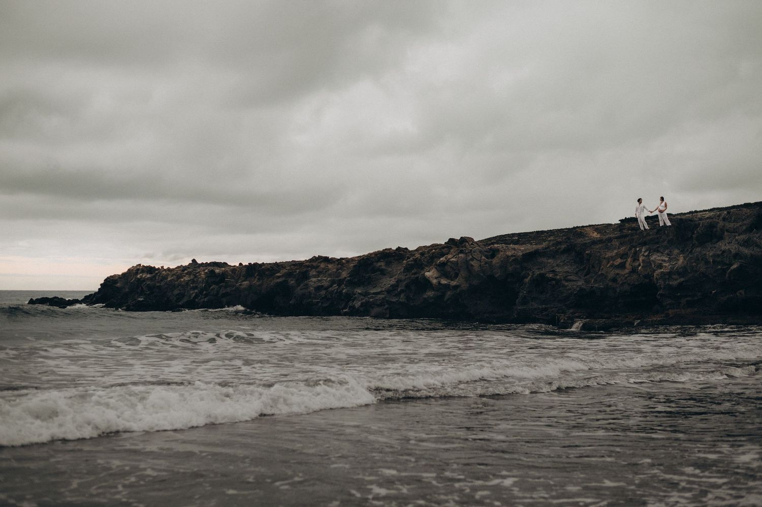 Two people stand on rocky coastal cliffs overlooking moody ocean waves under a dramatic overcast sky.