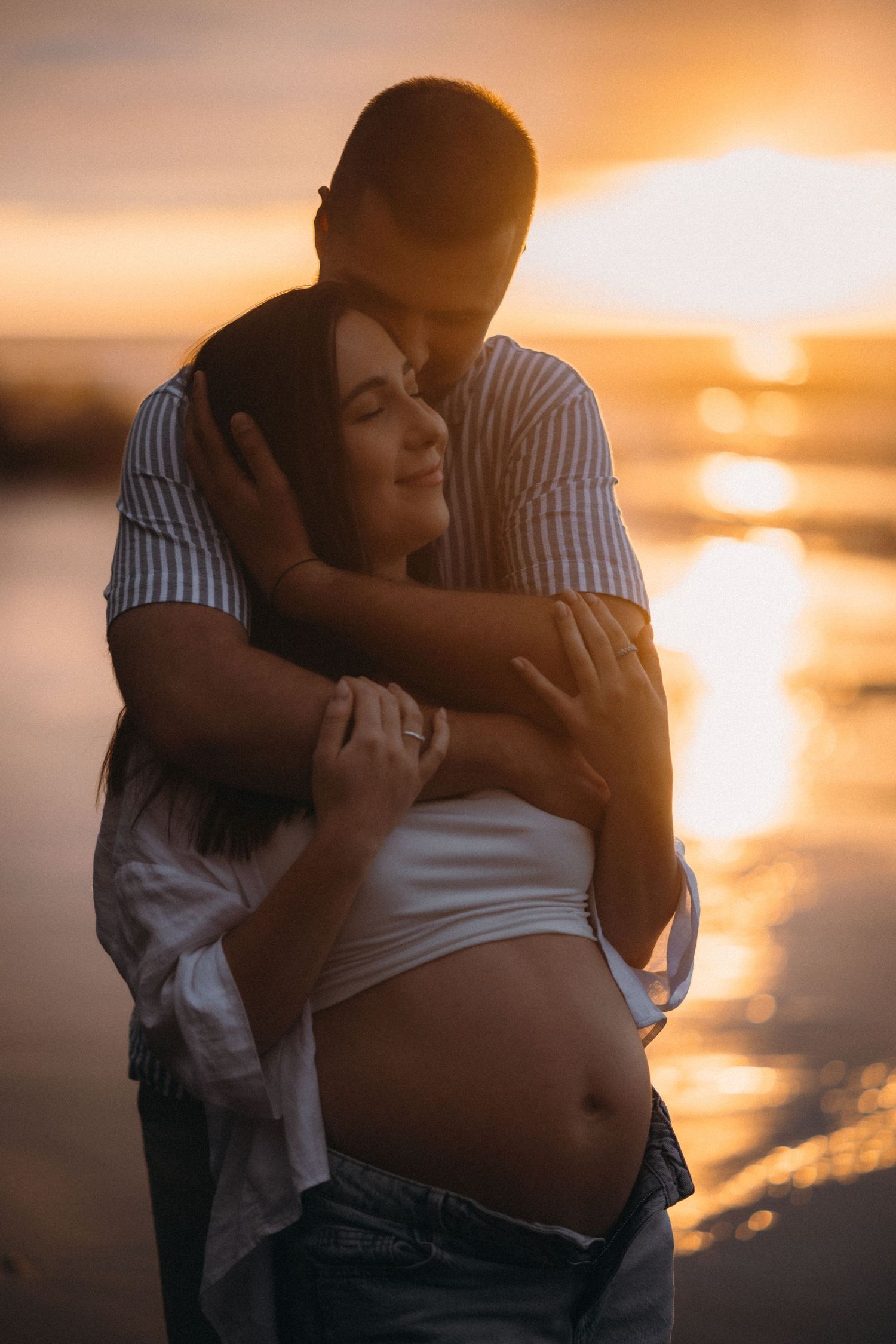 A couple embraces on a beach at golden sunset, the woman visibly pregnant, both smiling peacefully near reflective water.