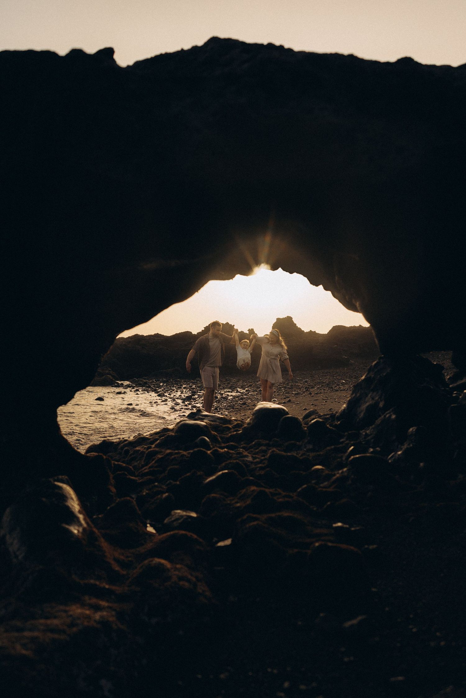 Couple silhouetted against golden sunset light framed by rocky sea cave arch on a pebbled beach.
