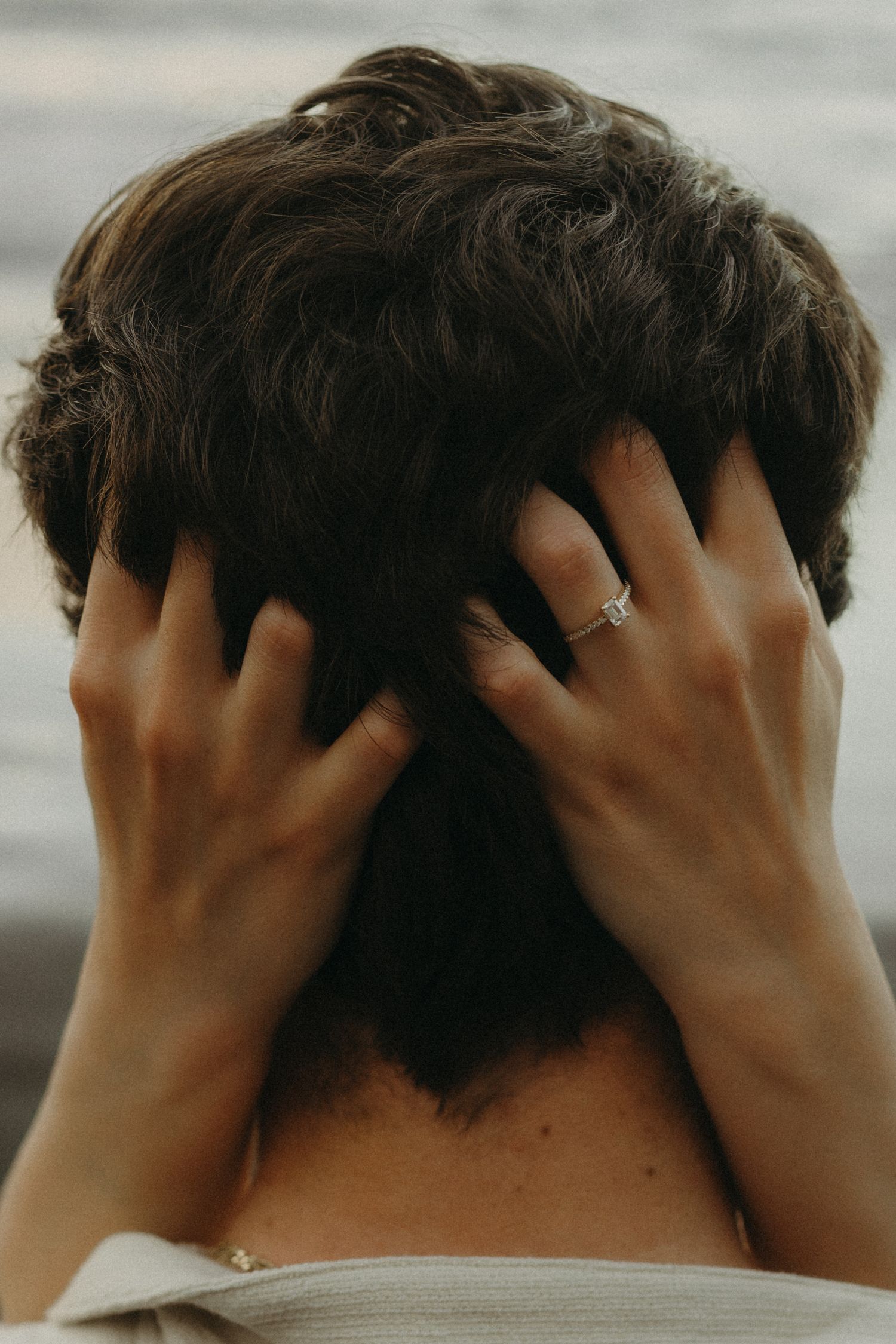 Person with dark curly hair seen from behind, hands with engagement ring gently cradling their head.