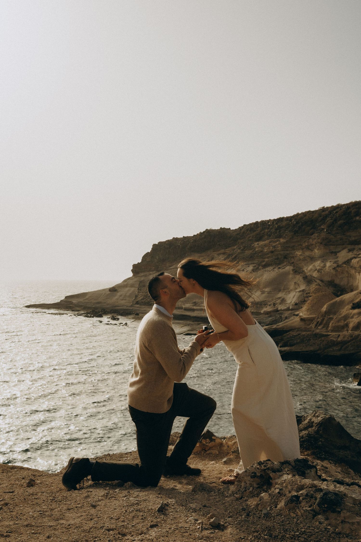 Surprise proposal in Costa Adeje, Tenerife on rocky coastal cliffs with a romantic kiss at sunset.
