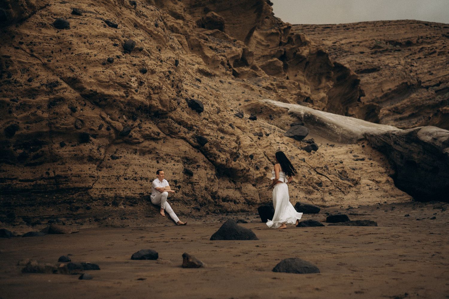 Couple in white attire poses on sandy beach against dramatic rocky cliffs for a romantic photoshoot.