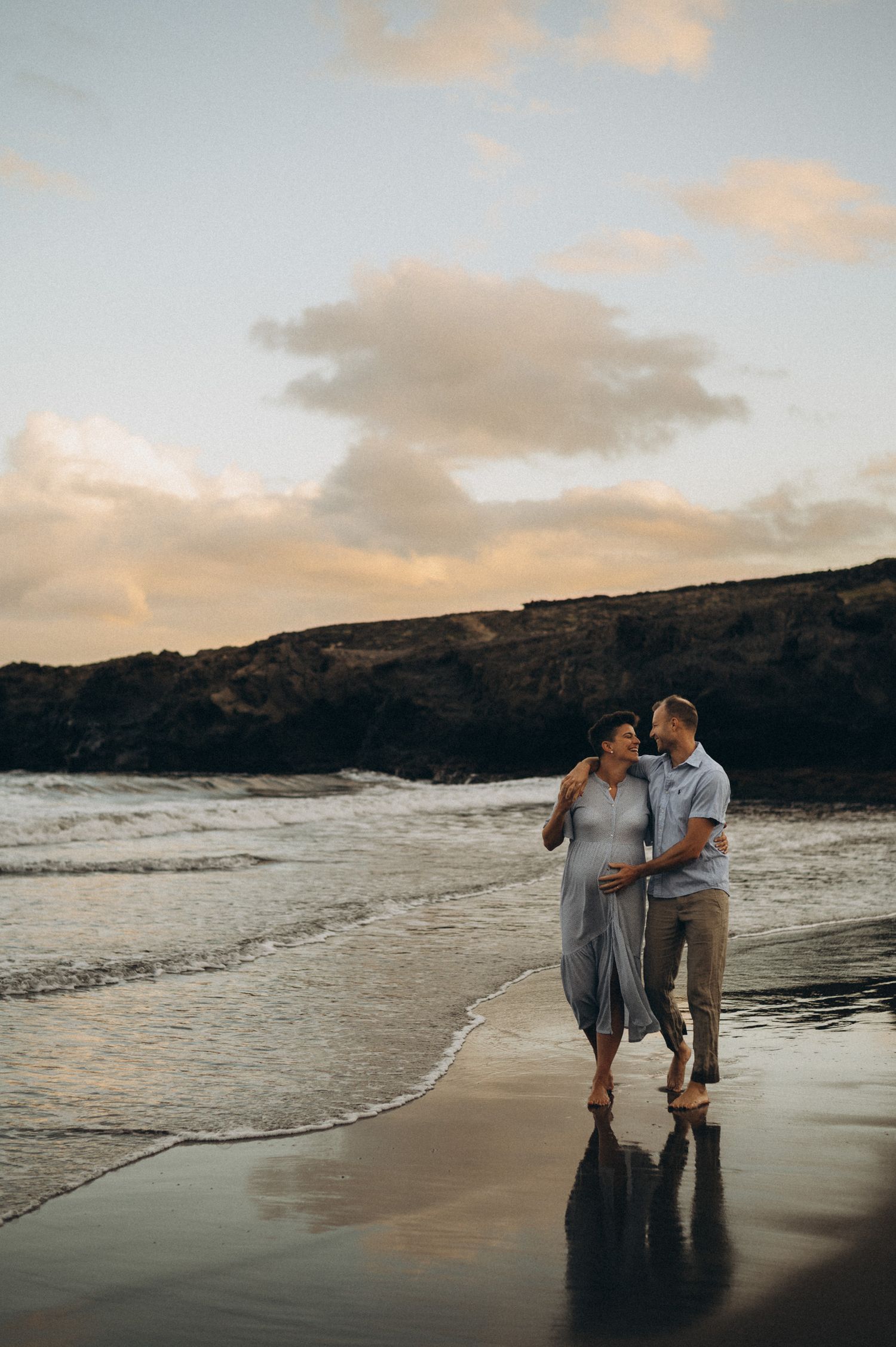 Couple walking on beach at sunset, woman in blue dress with baby bump, man in casual attire, waves and rocky cliffs behind.